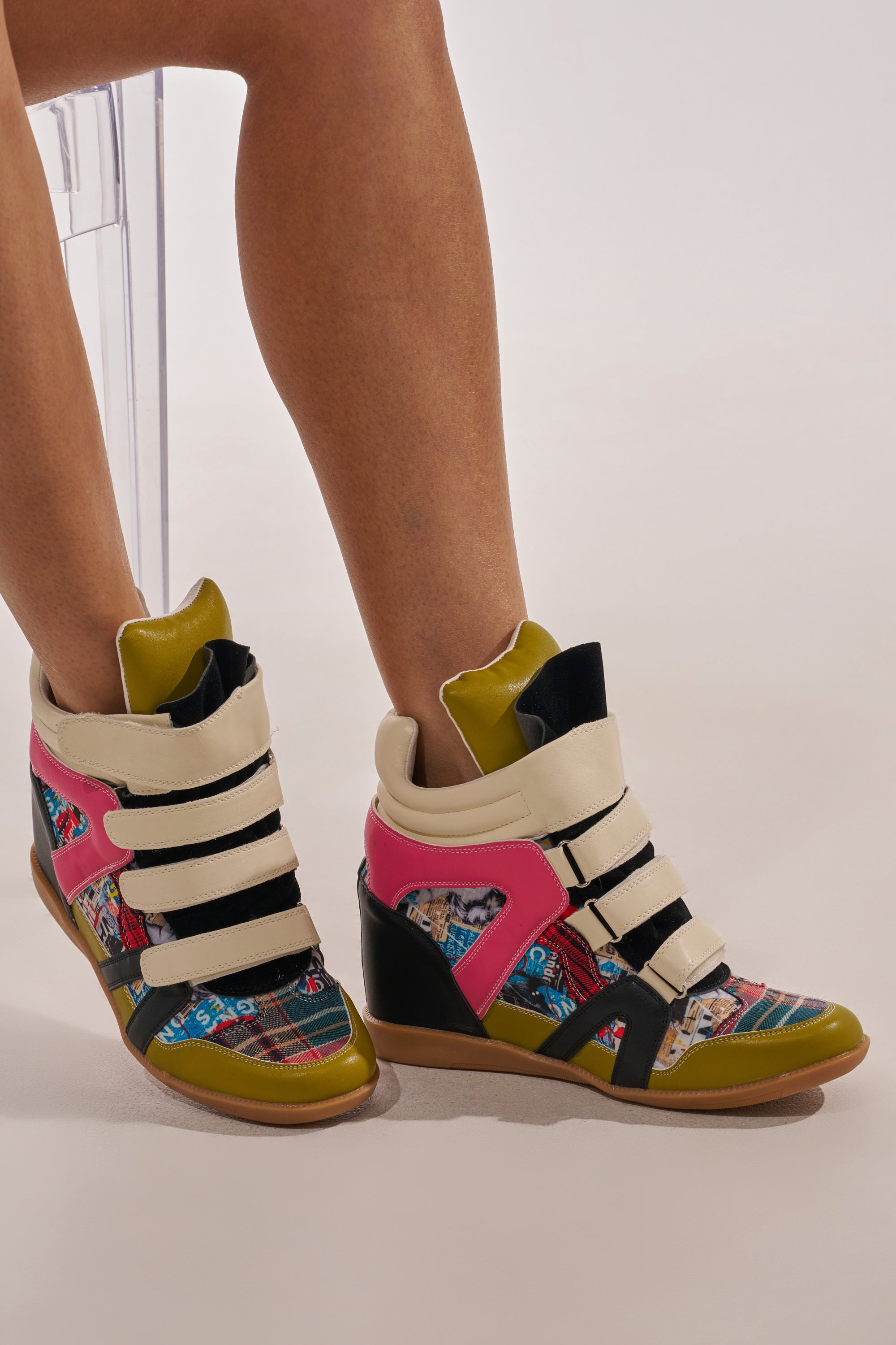 A person seated on a clear chair wears the AZALEA WANG BRYANT MULTI WEDGE SNEAKER—colorful, fashion-forward shoes with multiple Velcro straps and pink, black, cream, and multicolored patchwork accents. 0