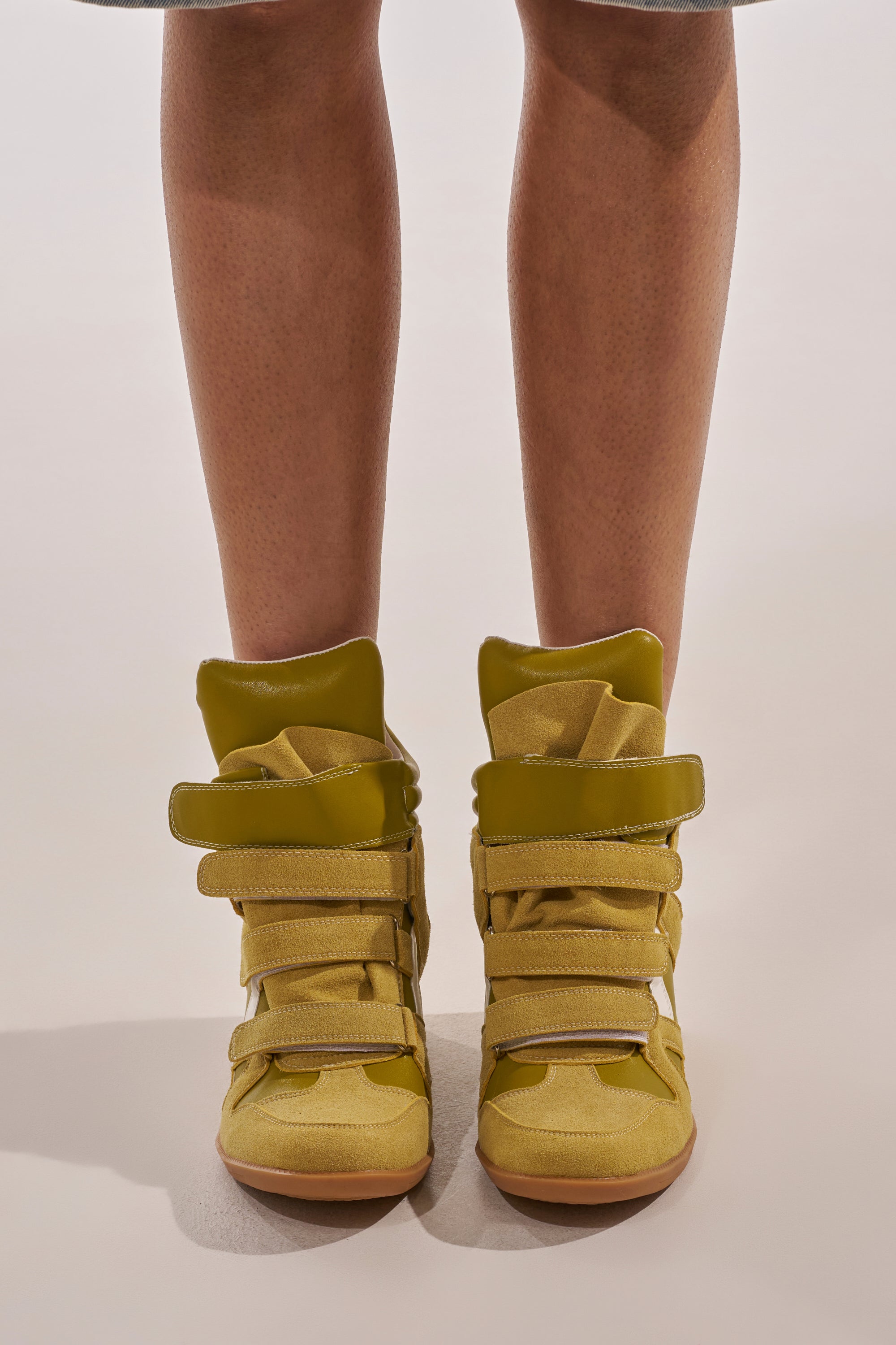 A person stands on a light surface wearing AZALEA WANG BRYANT OLIVE WEDGE SNEAKERS with thick straps and textured soles. Only the lower legs and feet are visible. 1