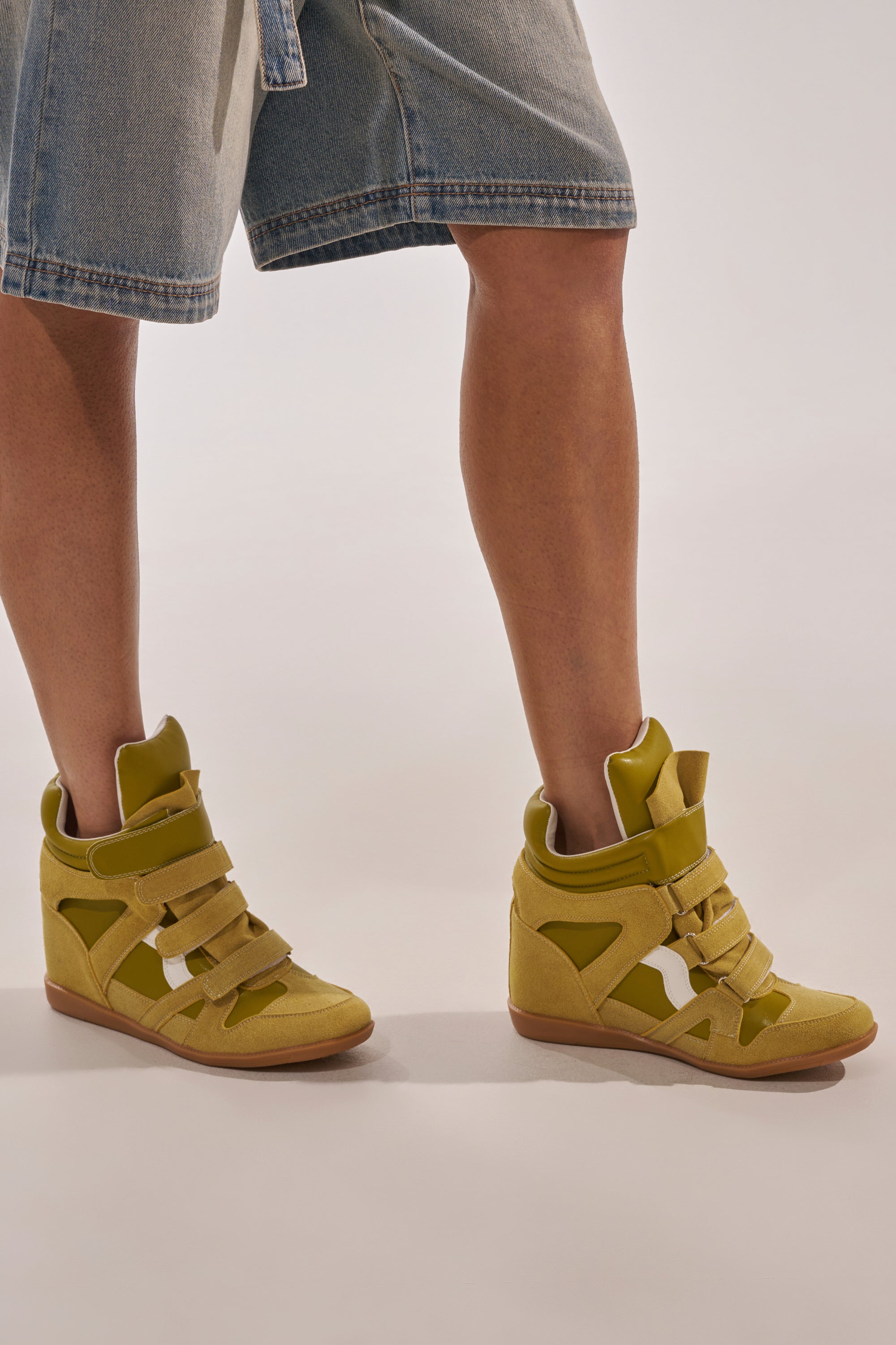 A person is shown from the knees down, wearing knee-length denim shorts and AZALEA WANG BRYANT OLIVE WEDGE SNEAKERS with velcro straps, standing against a neutral background. 2