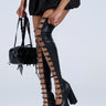 A person is wearing AZALEA WANG BRYNNE black chunky platform boots with rhinestone buckles, holding a black faux leather handbag with ruffle details, and sporting a short black skirt against a plain backdrop.