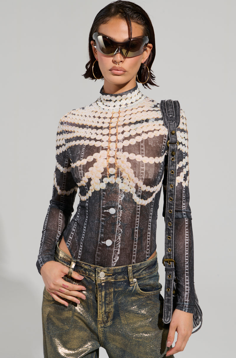 A person with short hair wears futuristic, angular sunglasses and the LOCKED IN BODYSUIT adorned with pearl-like designs. Paired with metallic gold pants and a studded shoulder bag, they stand strikingly against a plain background, embodying avant-garde elegance.