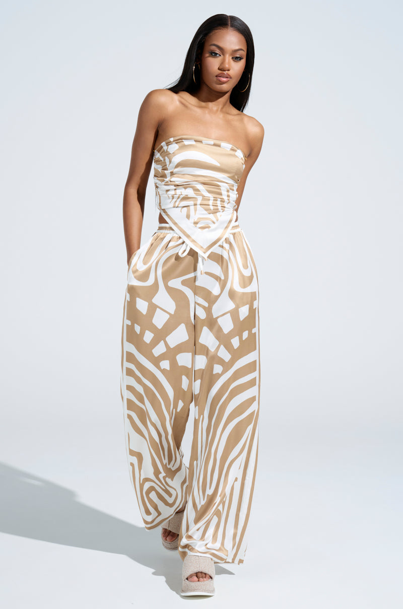 A woman confidently strides forward, her long hair flowing behind her, clad in a LOTS OF LOVE SATIN PALAZZO PANT featuring a high-rise fit with a geometric pattern in beige and white. The light plain background contrasts elegantly with the striking color block abstract print of her ensemble.
