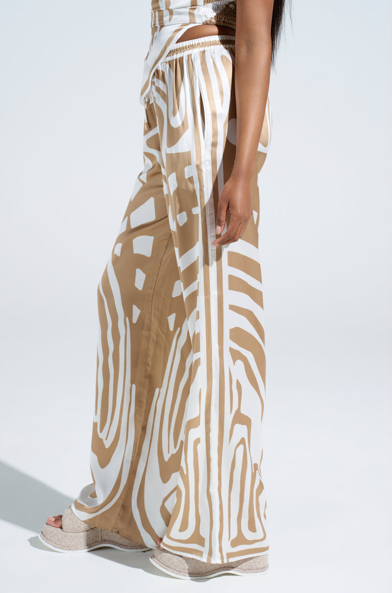 A person stands sideways wearing the LOTS OF LOVE SATIN PALAZZO PANT, featuring a tan and white abstract print with flowing, loose-fitting, high-rise design against a light gray background.