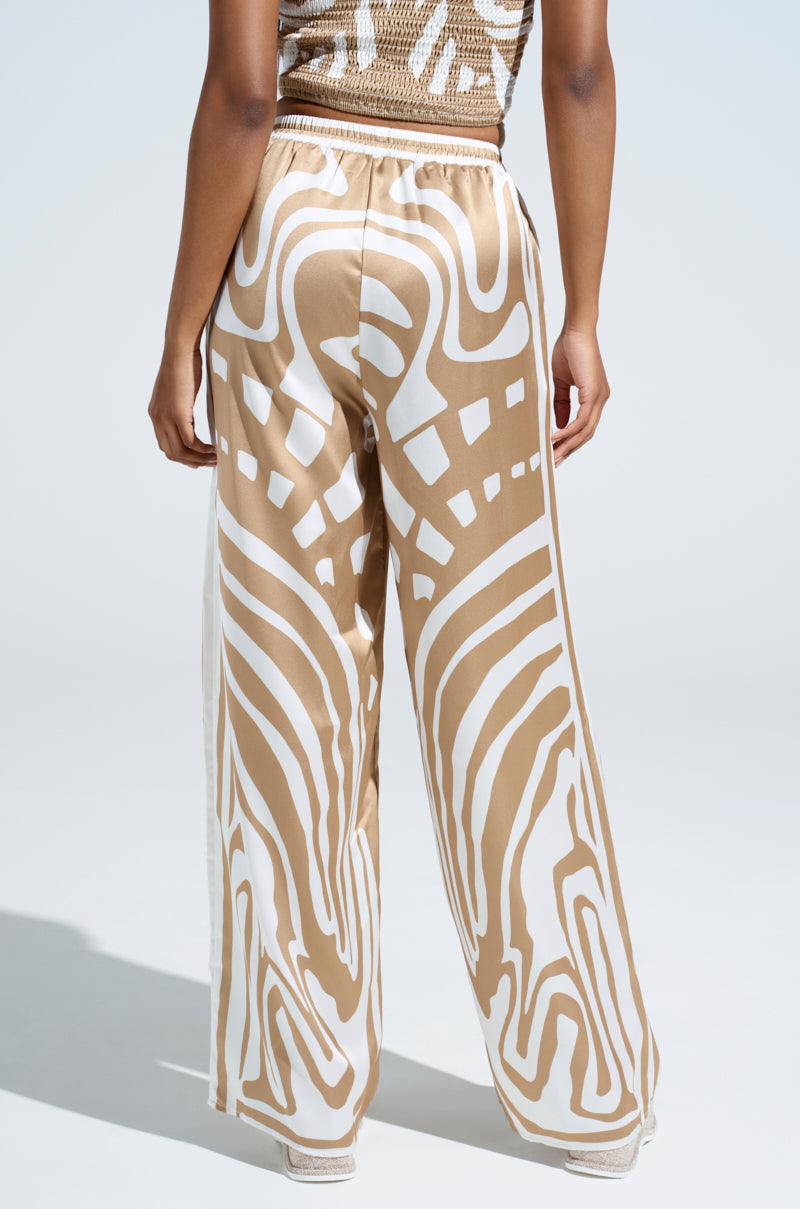 A person stands with their back to the camera, showcasing the LOTS OF LOVE SATIN PALAZZO PANT. These wide-legged pants feature a high-rise fit and an eye-catching color block abstract print in beige and white against a plain, light-colored backdrop.
