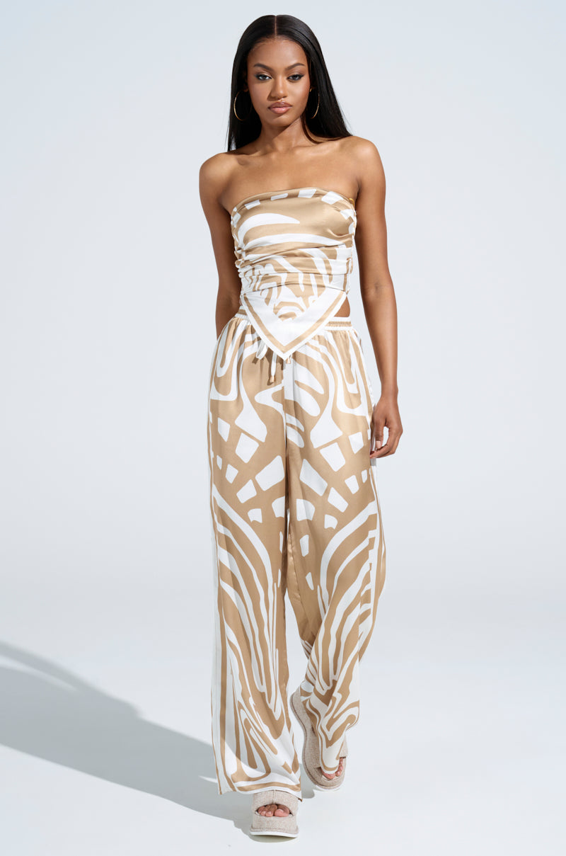 A woman in a LOTS OF LOVE SATIN PALAZZO PANT with abstract print, beige and white hues, walks gracefully and confidently. Her long hair flows past hoop earrings, paired with platform sandals. The high-rise fit adds elegance against the plain, light-colored background.