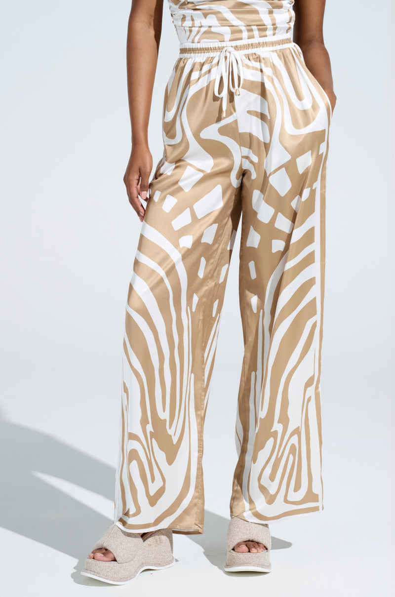 Wearing the LOTS OF LOVE SATIN PALAZZO PANT, the person exudes confidence in high-rise, beige and white wide-leg pants with a drawstring waist. Open-toed platform sandals complete the look against a light gray backdrop, enhancing their elegance.