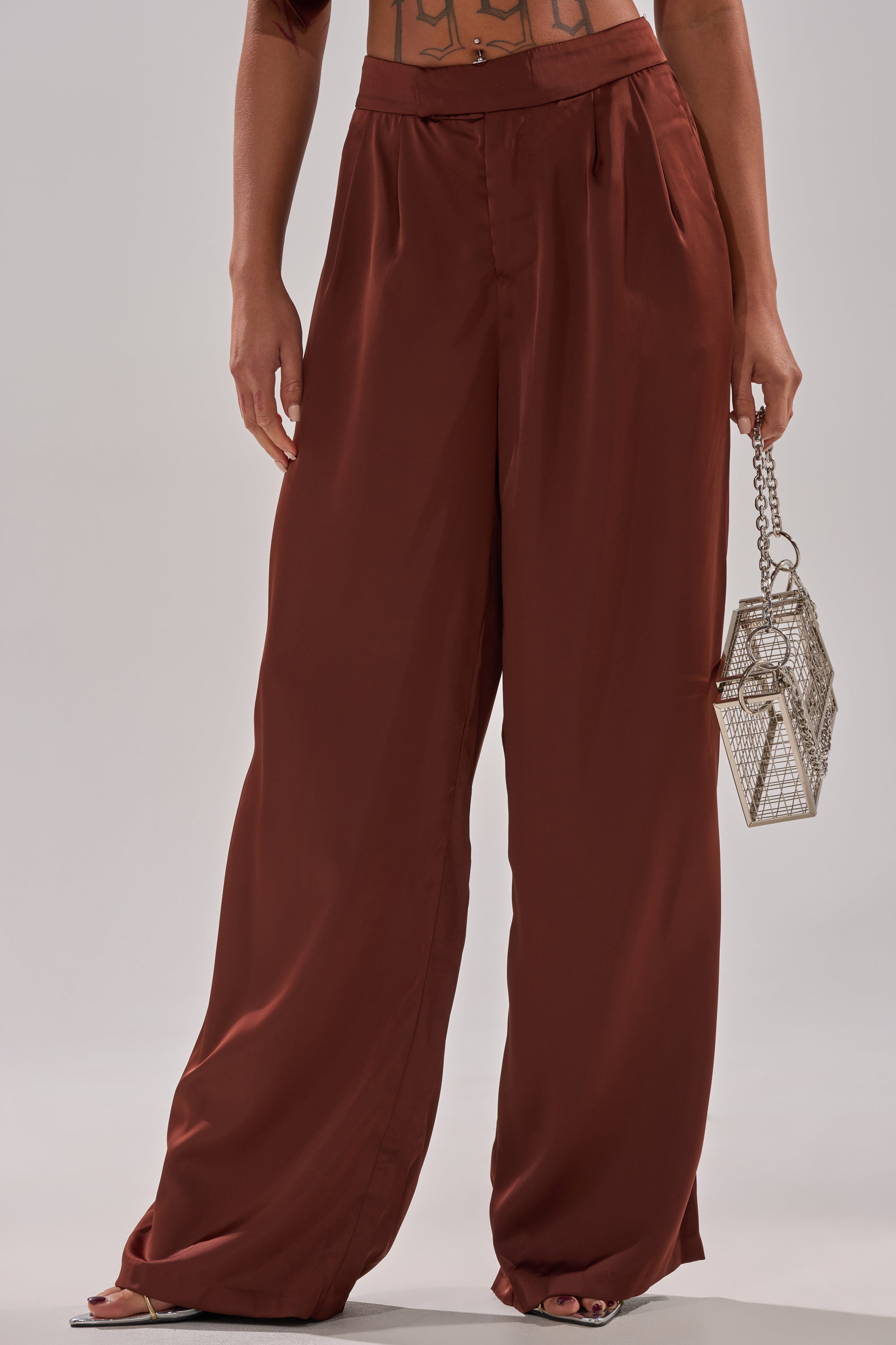 A person wears rust-colored satin HIGH SOCIETY TROUSER, designed as high-rise wide-leg pants, and holds a geometric silver handbag. Their midriff and hands are visible against a plain light background.