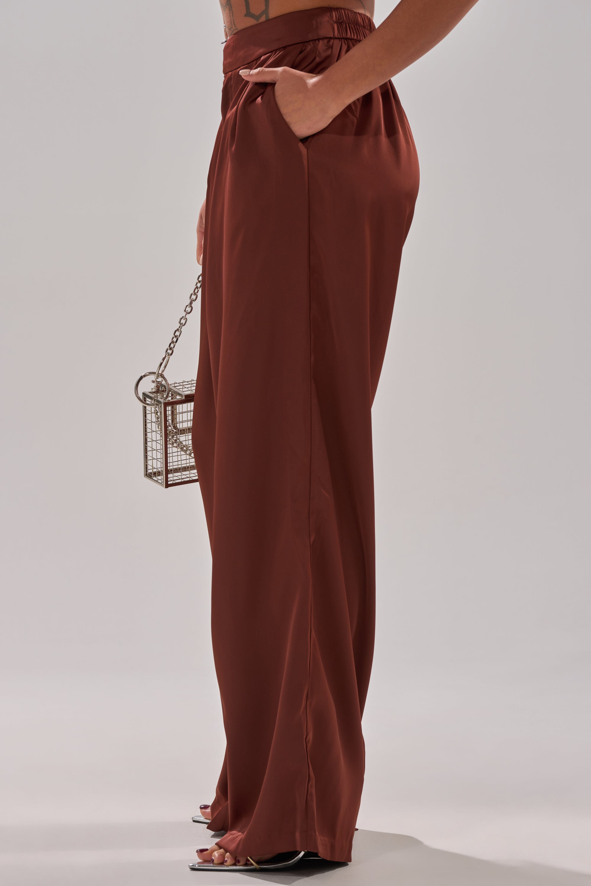 A person wears rust-colored HIGH SOCIETY TROUSER, high-waisted and wide-leg, with one hand in a pocket holding a metallic box purse. Only their lower half is shown against a light background.