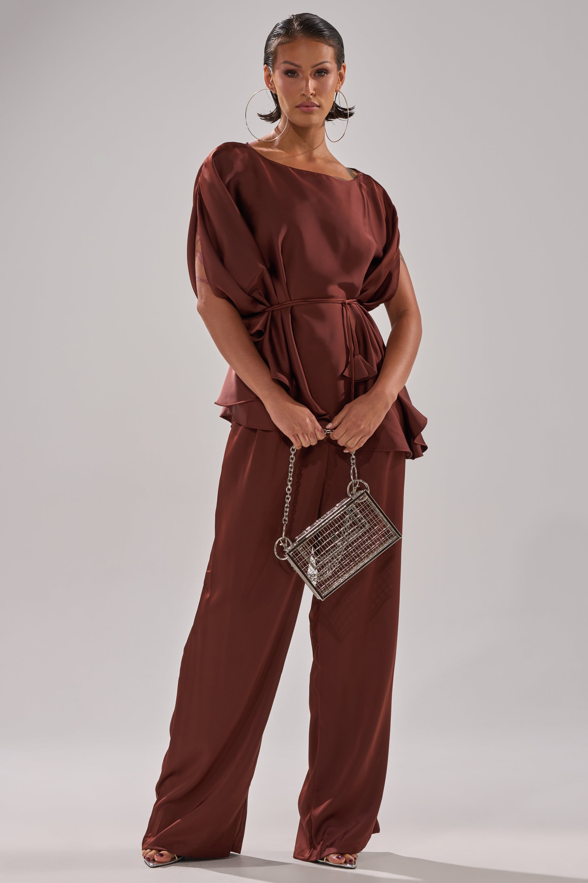 A woman poses in loose, chocolate-brown satin blouse and HIGH SOCIETY TROUSER, holding a small silver metallic purse with a chain strap. She accessorizes with large hoop earrings and slicked-back hair.