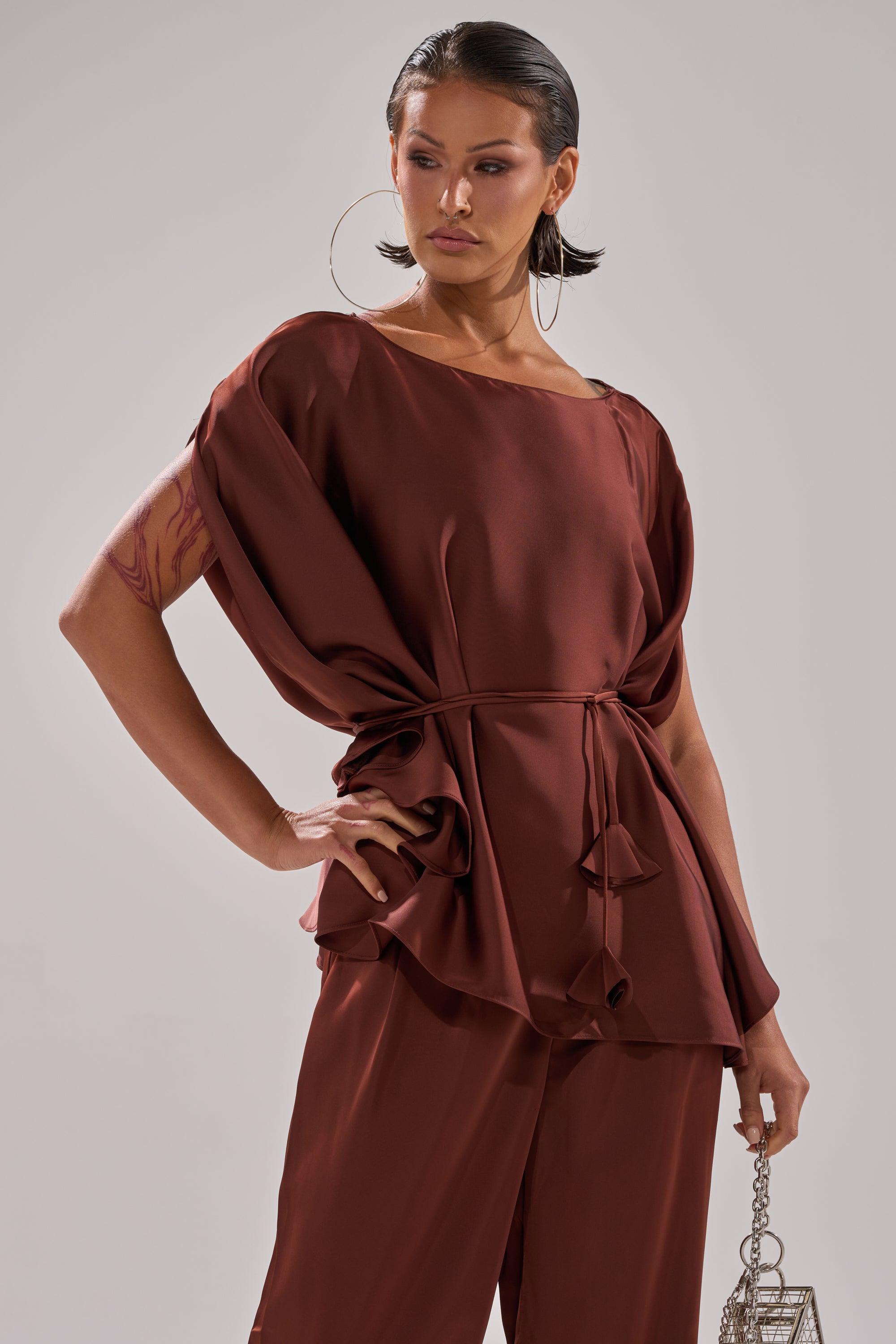 A woman wears the HIGH SOCIETY BLOUSE in dark brown satin with batwing sleeves, styled with matching pants. She finishes her chic look with large hoop earrings, an upper arm tattoo, and a small metallic handbag.