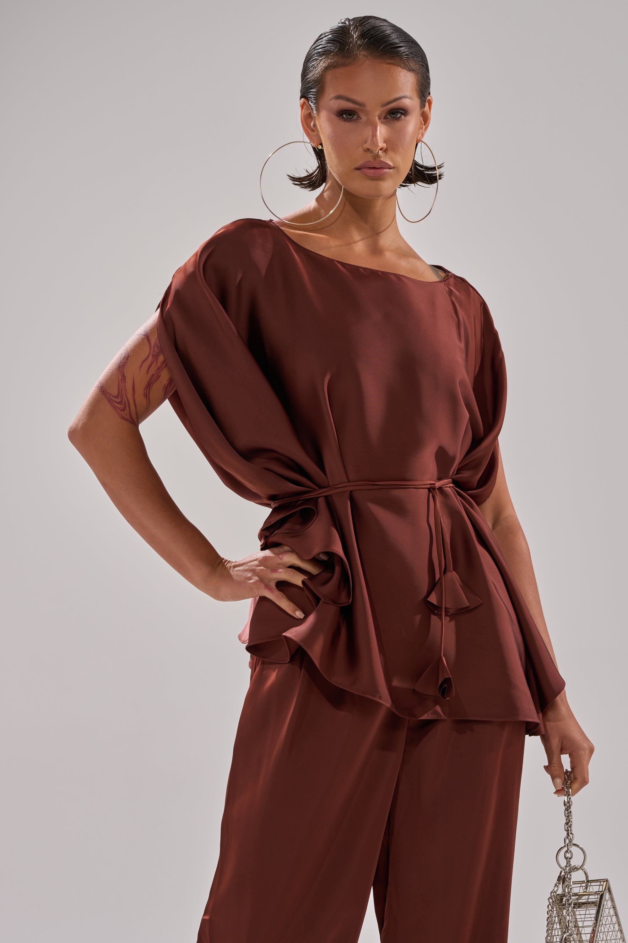 A woman confidently poses in the HIGH SOCIETY BLOUSE— a loose-fitting brown satin top with batwing sleeves, paired with matching pants. She accessorizes with large hoop earrings and a small silver handbag, showcasing her arm tattoos.