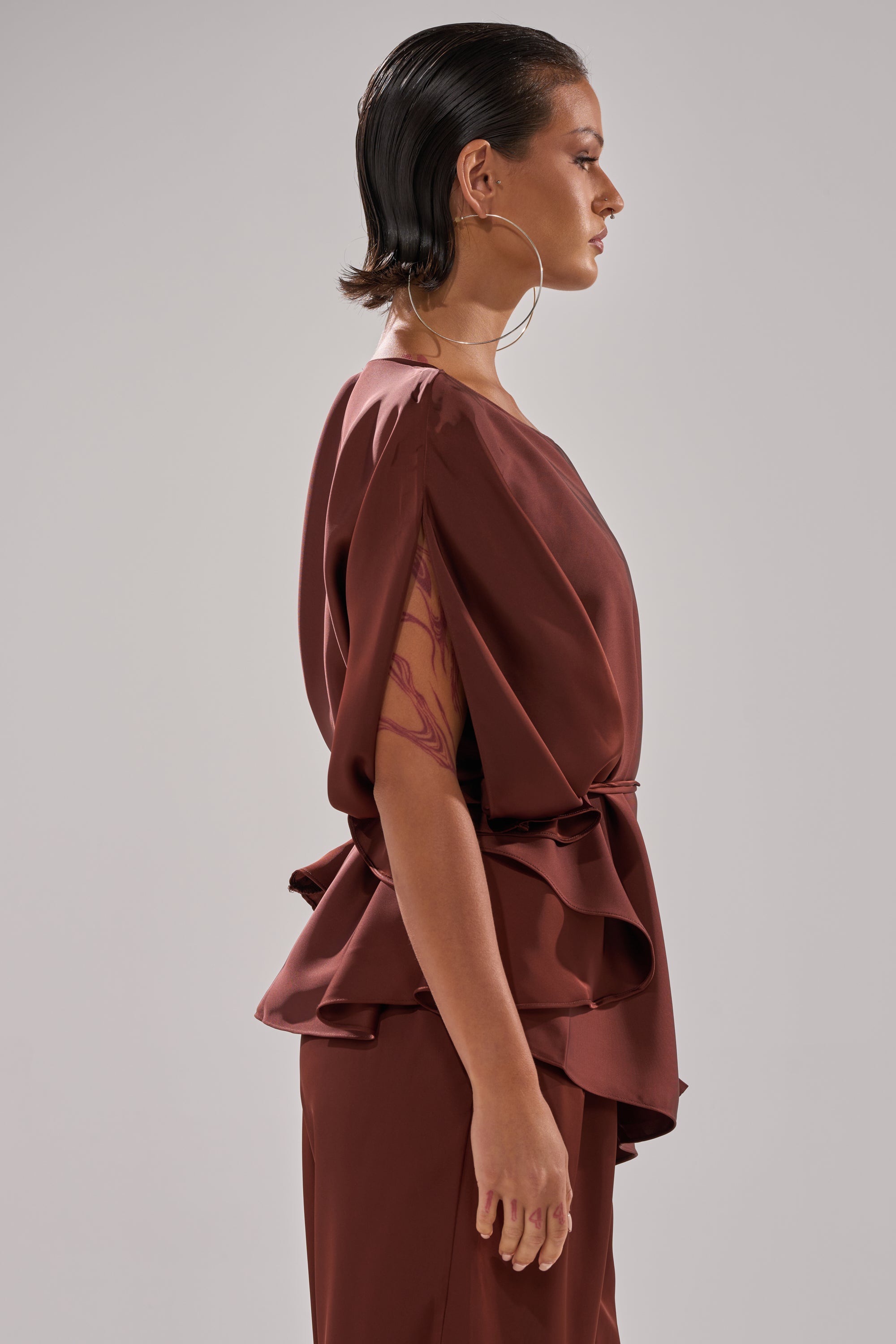A person with slicked-back dark hair models the loose, ruffled HIGH SOCIETY BLOUSE in brown with matching pants, a large hoop earring, and an upper arm tattoo, standing before a plain light gray background.