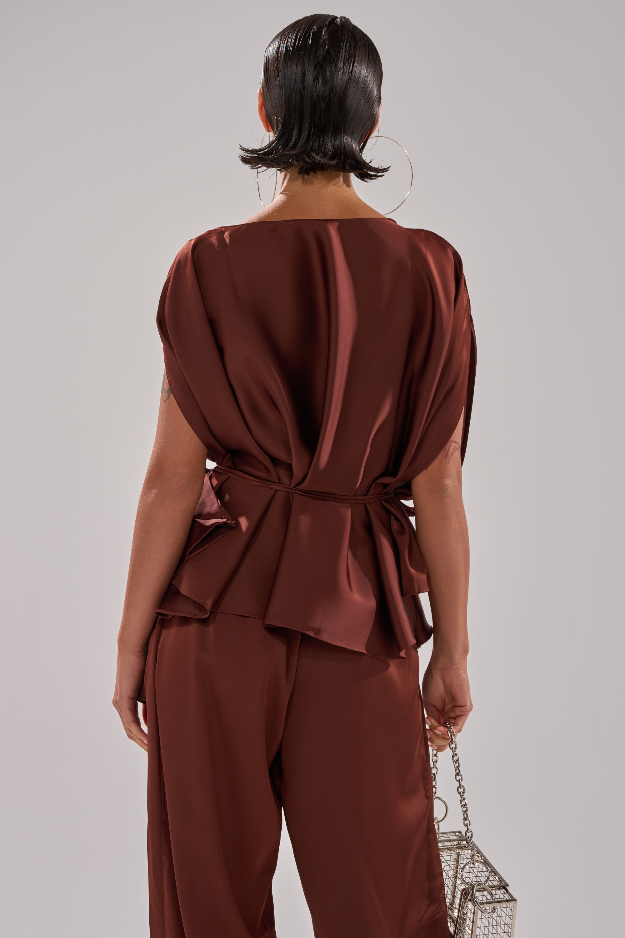 Seen from behind, a person with short, slicked-back hair wears the HIGH SOCIETY BLOUSE in deep brown satin with matching pants, accessorized with a metallic geometric purse and bold hoop earrings.