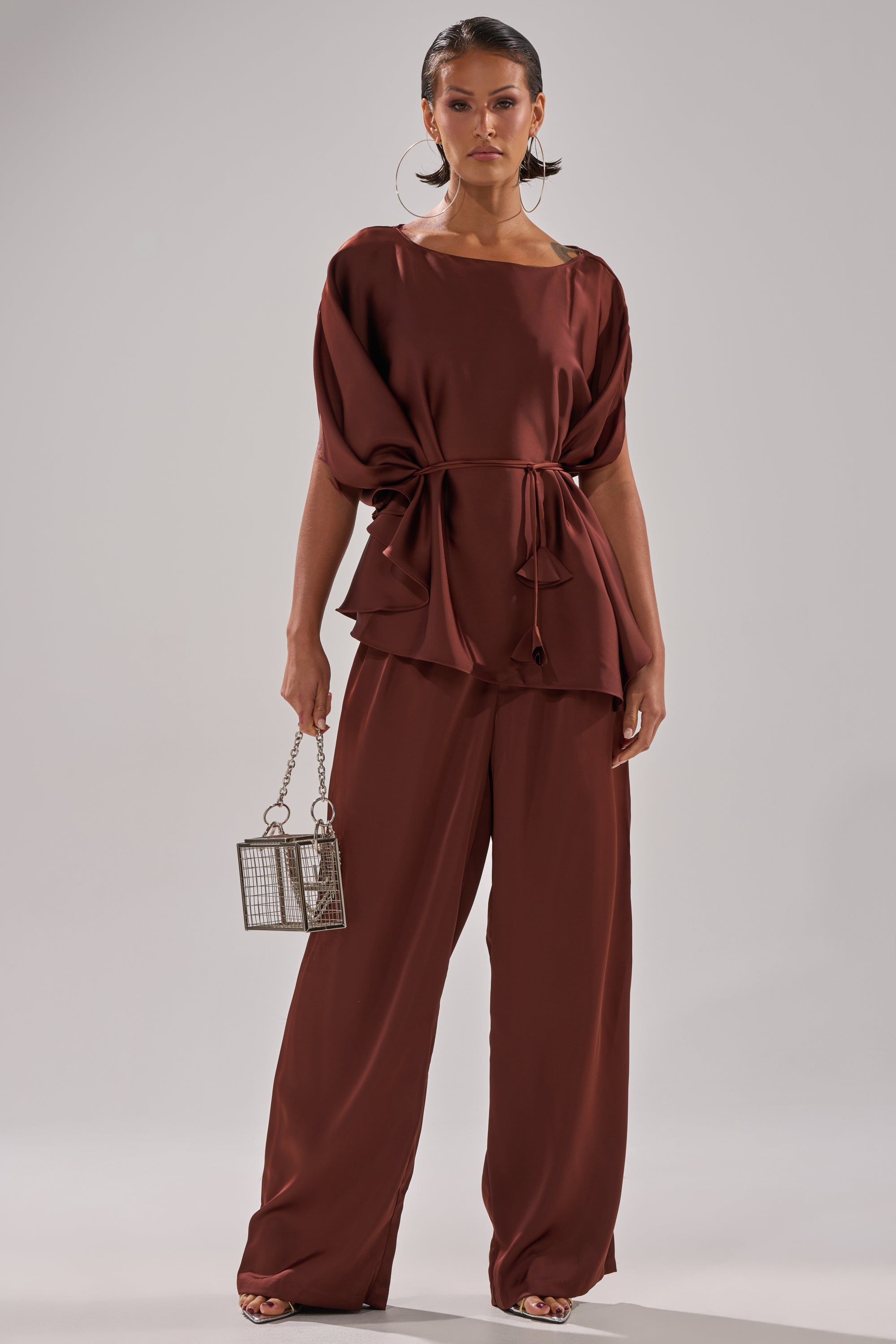 A woman wears the rust-colored HIGH SOCIETY BLOUSE with batwing sleeves and a waist tie, matching wide-leg pants, large hoop earrings, and carries a small metallic box handbag.