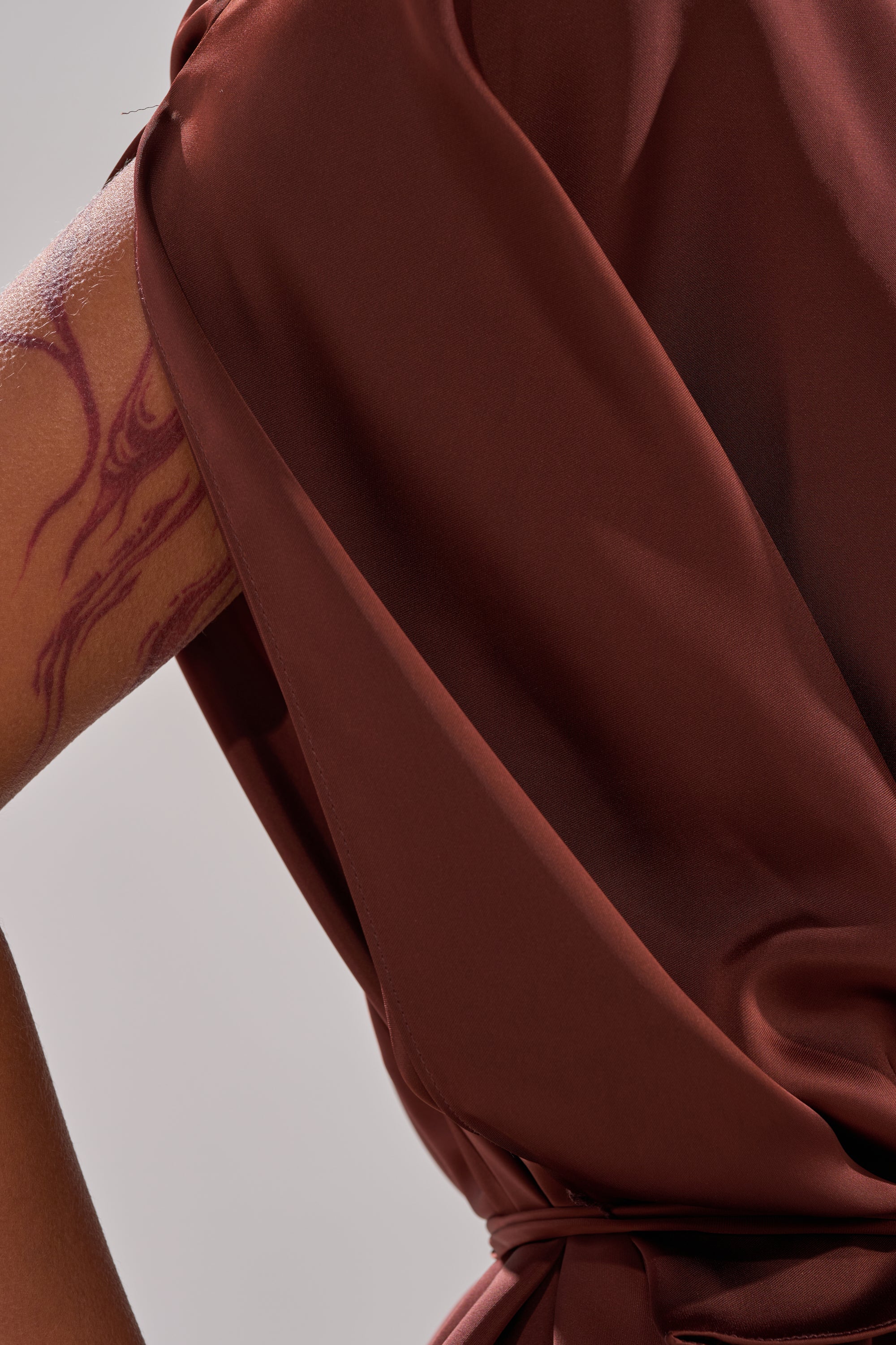 A close-up shows someone wearing a silky brown HIGH SOCIETY BLOUSE with batwing sleeves. Part of their arm, featuring a bold abstract tattoo, is visible against a simple neutral background.