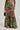 A person is wearing the "ON VACAY WIDE LEG PANT IN GREEN MULTI," a pair of colorful, high-waisted, wide-leg pants featuring a vibrant, eclectic pattern with shades of green, pink, purple, and orange. These tropical-themed pants have a tiered and ruffled design. They are also holding a black handbag, with their shoes peeking out at the bottom.