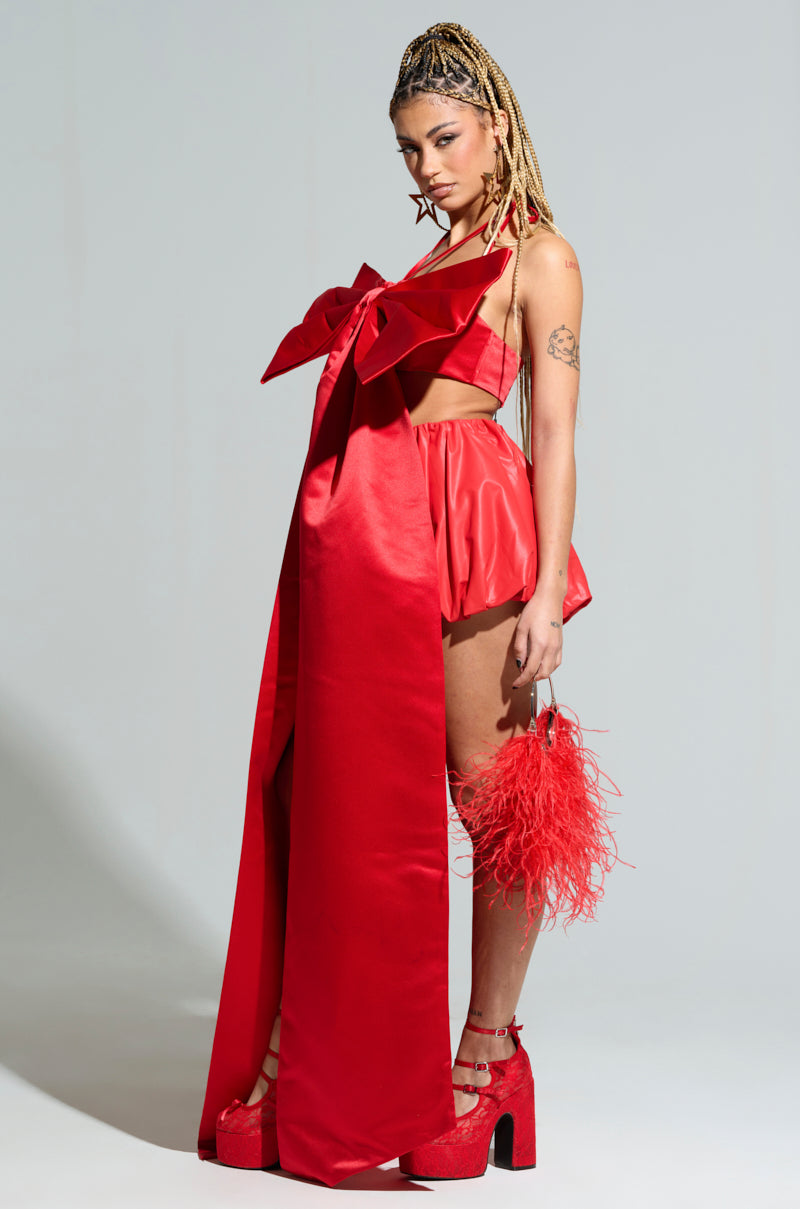 Dressed in the eye-catching MILA BUBBLE SKIRT with a large bow and flowing fabric, a person exudes confidence. They hold a red feathered accessory and are wearing red platform heels. Their long braided hair completes the striking ensemble.