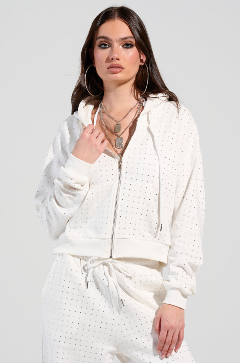 A person with long dark hair is wearing a white AKIRA Label Bubbles Rhinestone Embellished Zip-Up Sweatshirt and dotted pattern pants. They are accessorized with large hoop earrings and layered necklaces, exuding style in their stretchy cotton outfit. They stand in a neutral pose against a plain background.