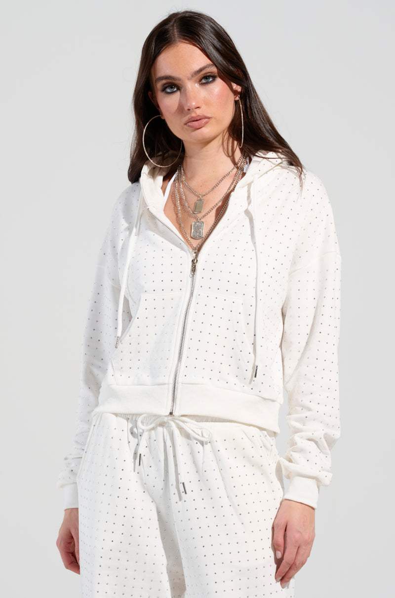 A woman with long dark hair is wearing the BUBBLES Rhinestone Embellished Zip Up Sweatshirt in White, featuring a design similar to small polka dots. She accessorizes with large hoop earrings and layered necklaces, maintaining a neutral expression as she stands against a plain background.