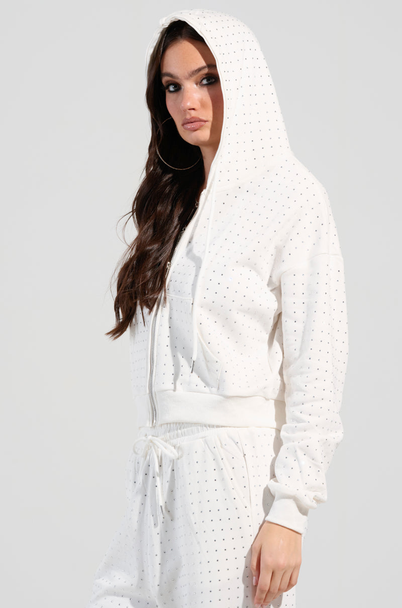 A person with long dark hair is wearing the BUBBLES Rhinestone Embellished Zip Up Sweatshirt in White from the AKIRA Label. The white hooded outfit, adorned with small dotted patterns, adds an elegant touch. They stand against a plain background, looking slightly to the side.