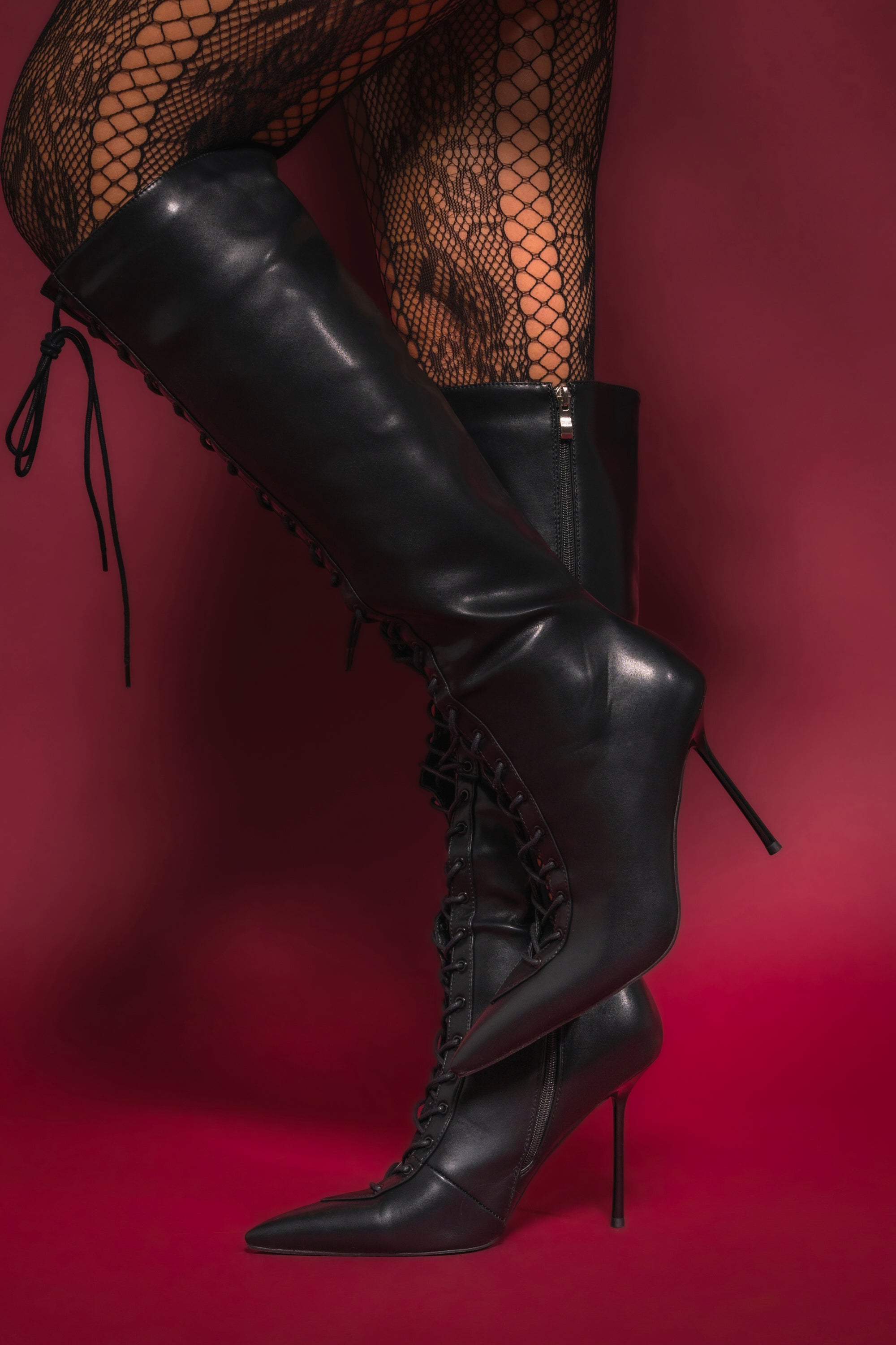 A person poses against a red background wearing black fishnet stockings and the AZALEA WANG BUCHANAN LACE UP BOOT IN BLACK, featuring pointed toes and stiletto heels. 0