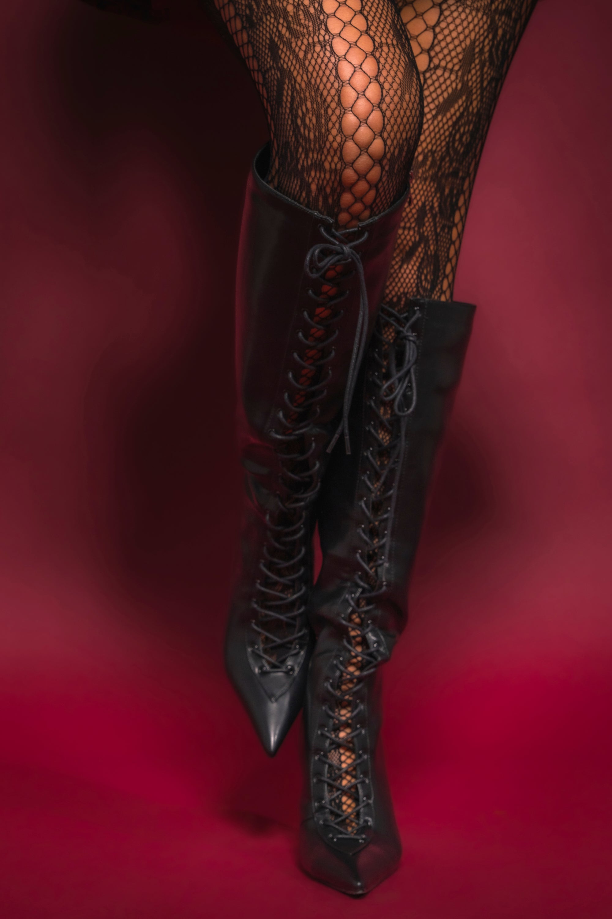 Model wearing fishnet stockings and the AZALEA WANG BUCHANAN LACE UP BOOT IN BLACK, posing against a deep red background. 1