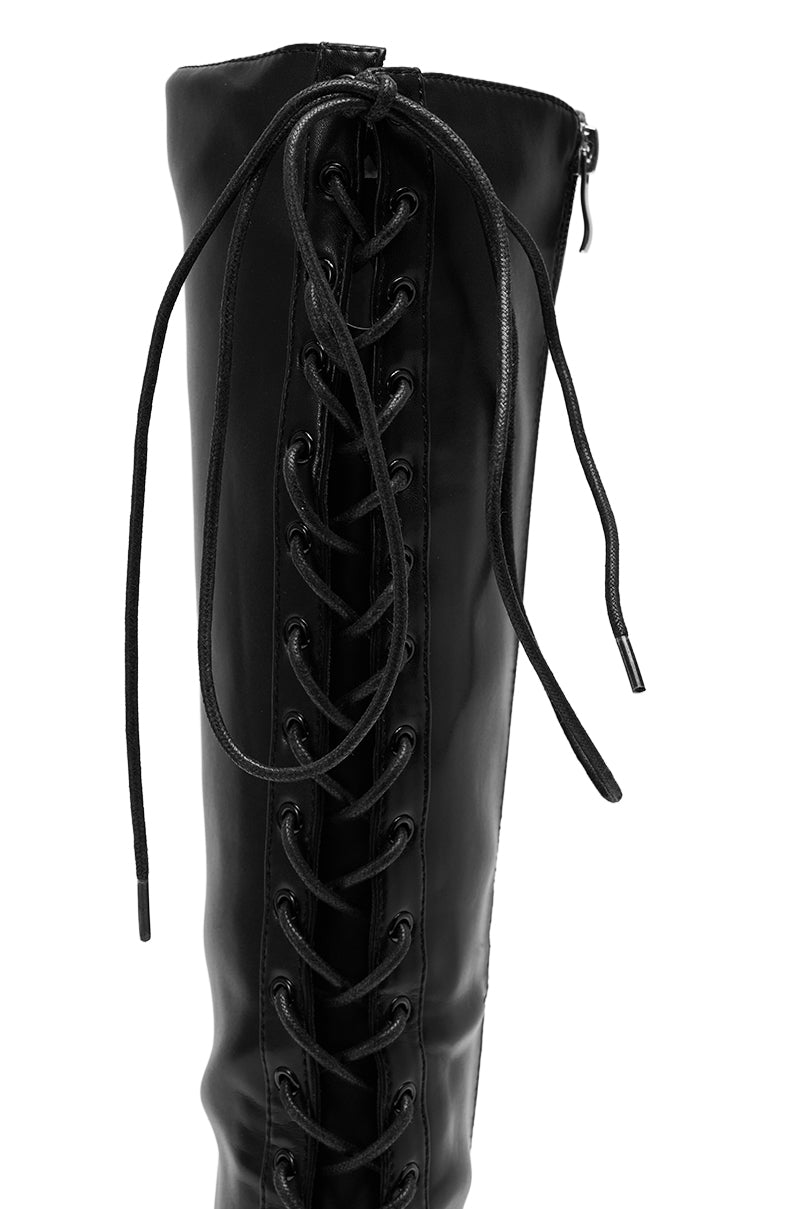 A close-up of the AZALEA WANG BUCHANAN LACE UP BOOT IN BLACK, a knee-high boot with front black laces, metal eyelets, a stiletto heel for height, and a partially visible side zipper at the top. 8