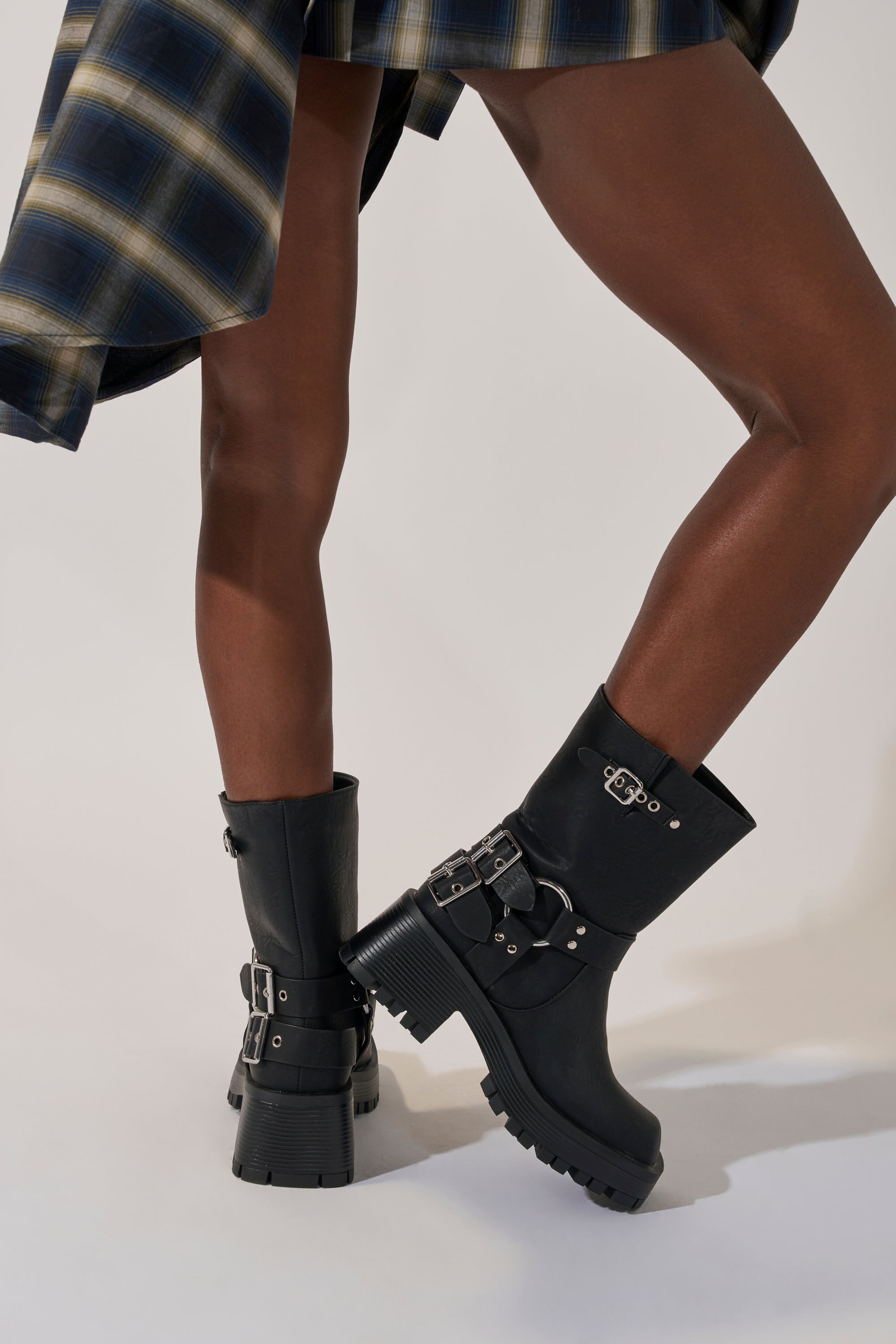 A person in a blue plaid skirt and AZALEA WANG BUCKED BLACK CHUNKY HEEL COMBAT BOOTIE stands with legs crossed against a light background.