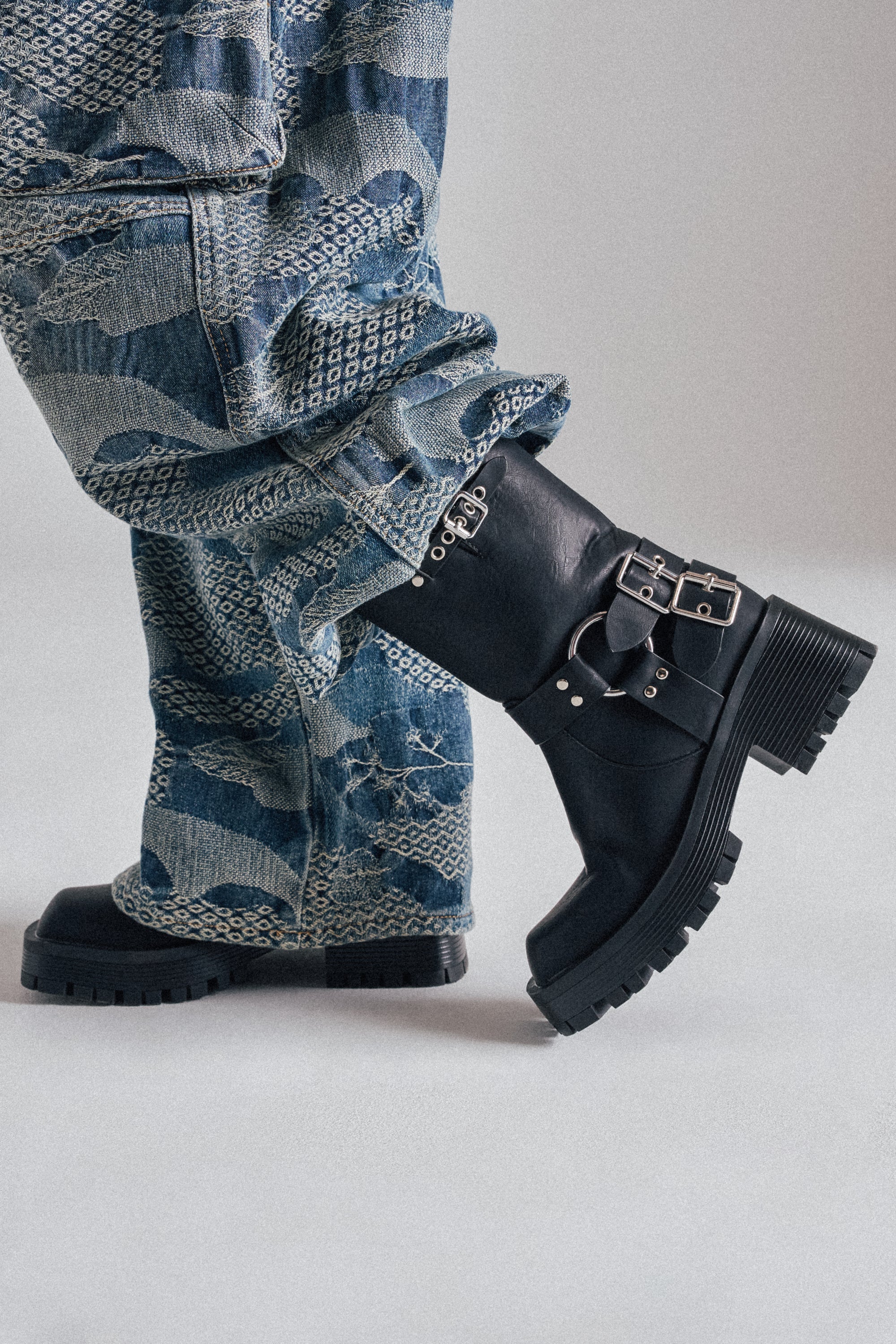 A person stands against a plain light background, wearing patterned blue jeans and AZALEA WANG BUCKED BLACK CHUNKY HEEL COMBAT BOOTIE. Only the lower legs are visible.