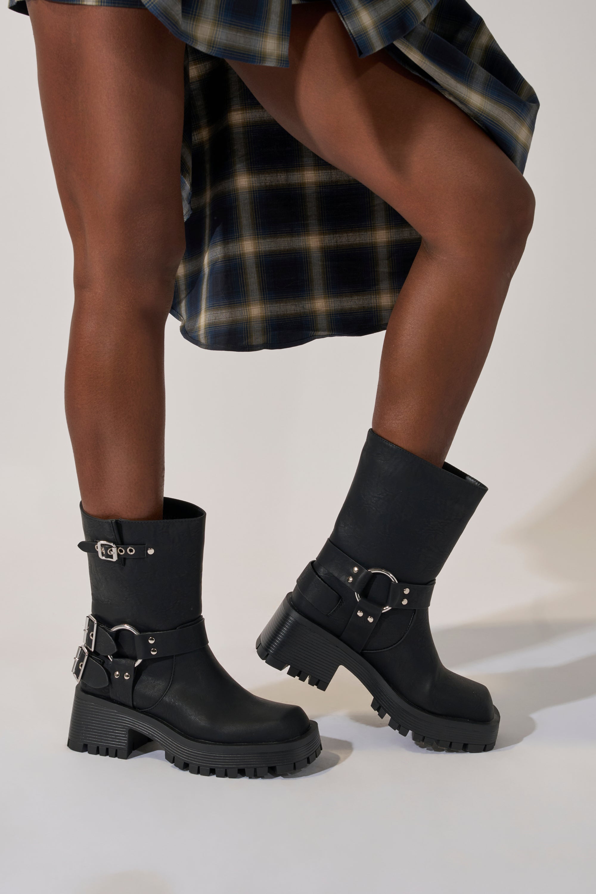 A person wearing a plaid skirt and AZALEA WANG BUCKED BLACK CHUNKY HEEL COMBAT BOOTIE with silver buckles stands on a light background; only their legs and the lower part of the skirt are visible.