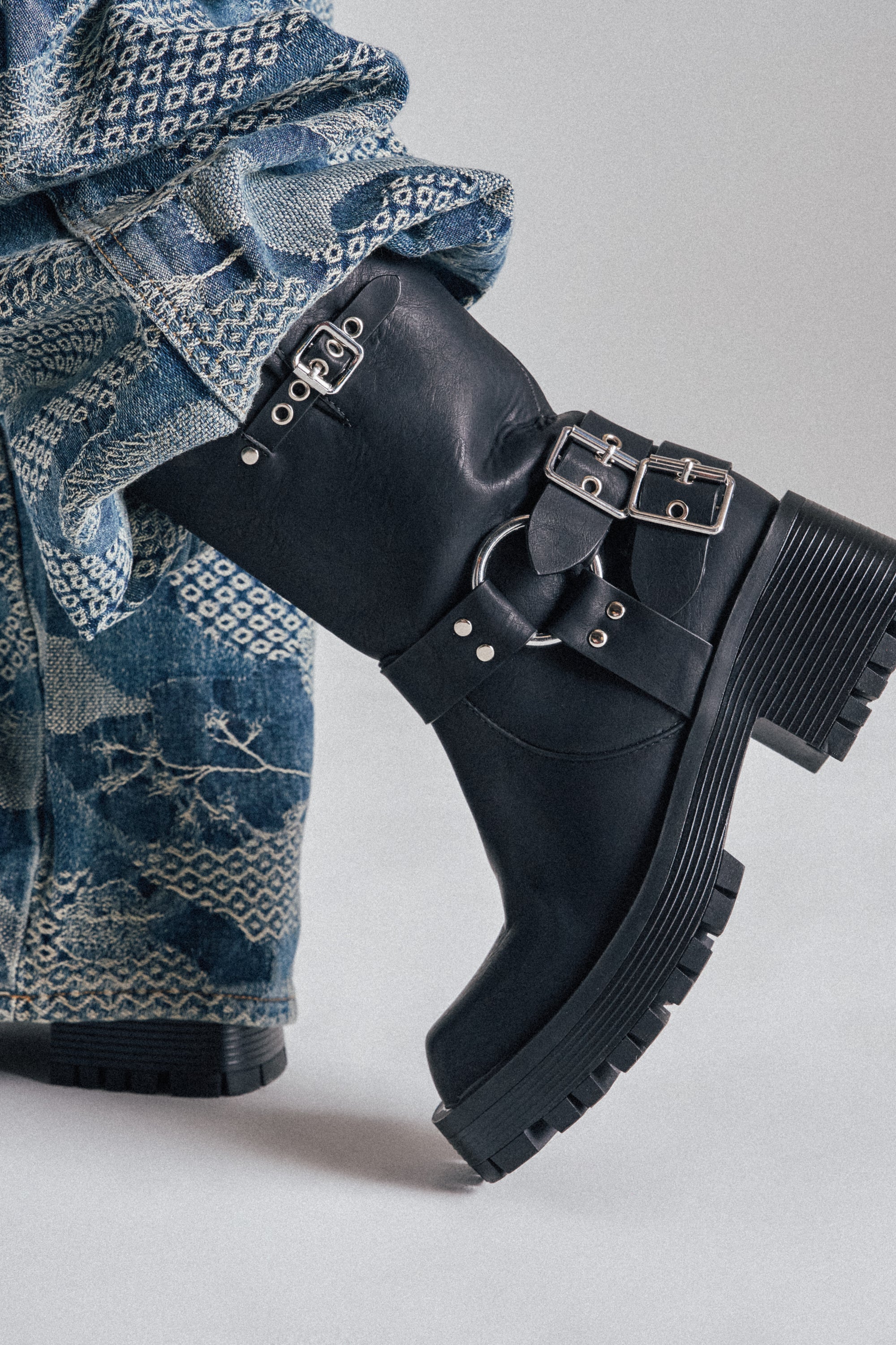 A close-up of a person wearing patterned blue jeans and AZALEA WANG BUCKED BLACK CHUNKY HEEL COMBAT BOOTIE with metal buckles and thick treaded soles, standing on a neutral gray background.