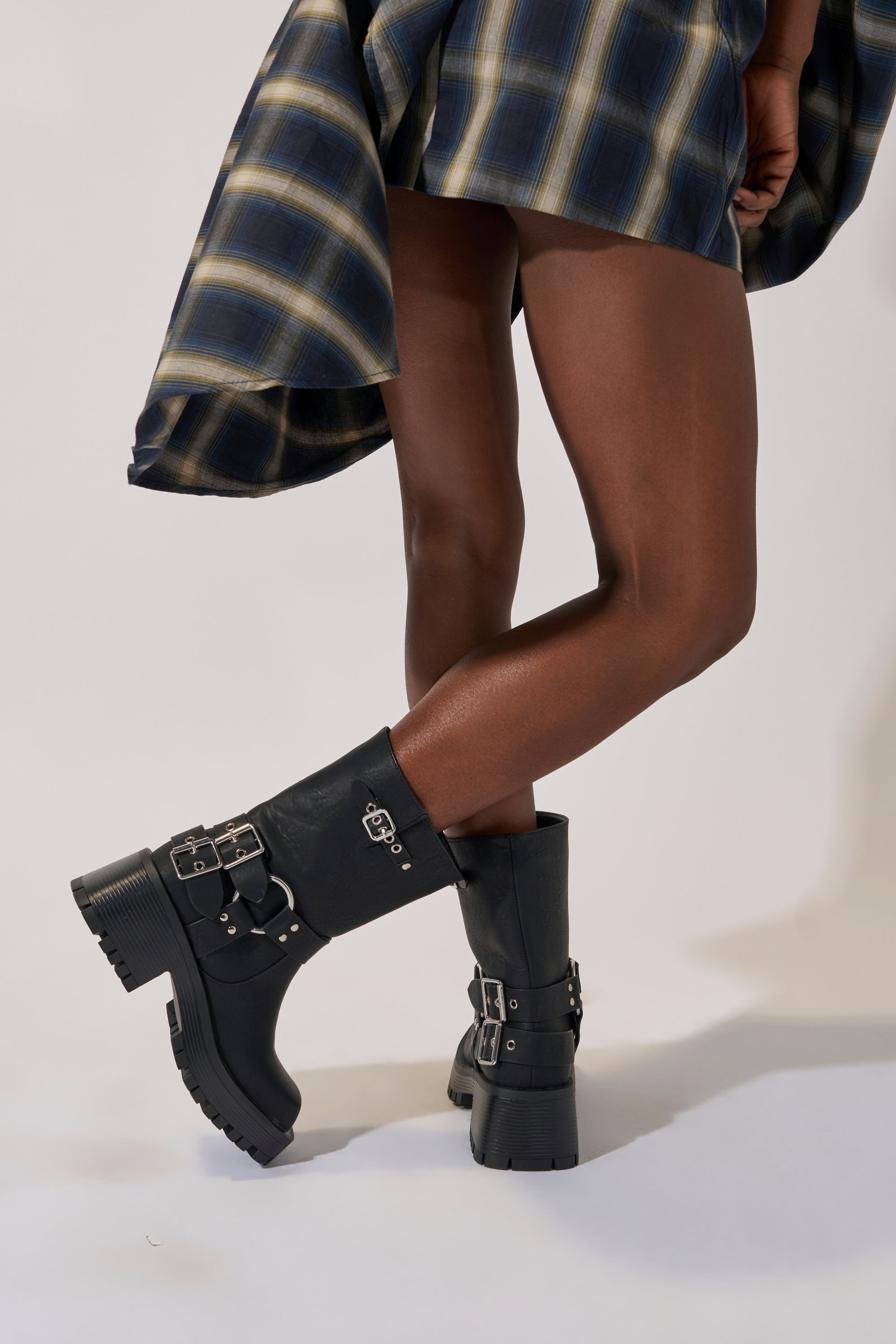 A person in a blue plaid skirt stands with legs crossed against a light background, wearing the AZALEA WANG BUCKED BLACK CHUNKY HEEL COMBAT BOOTIE.