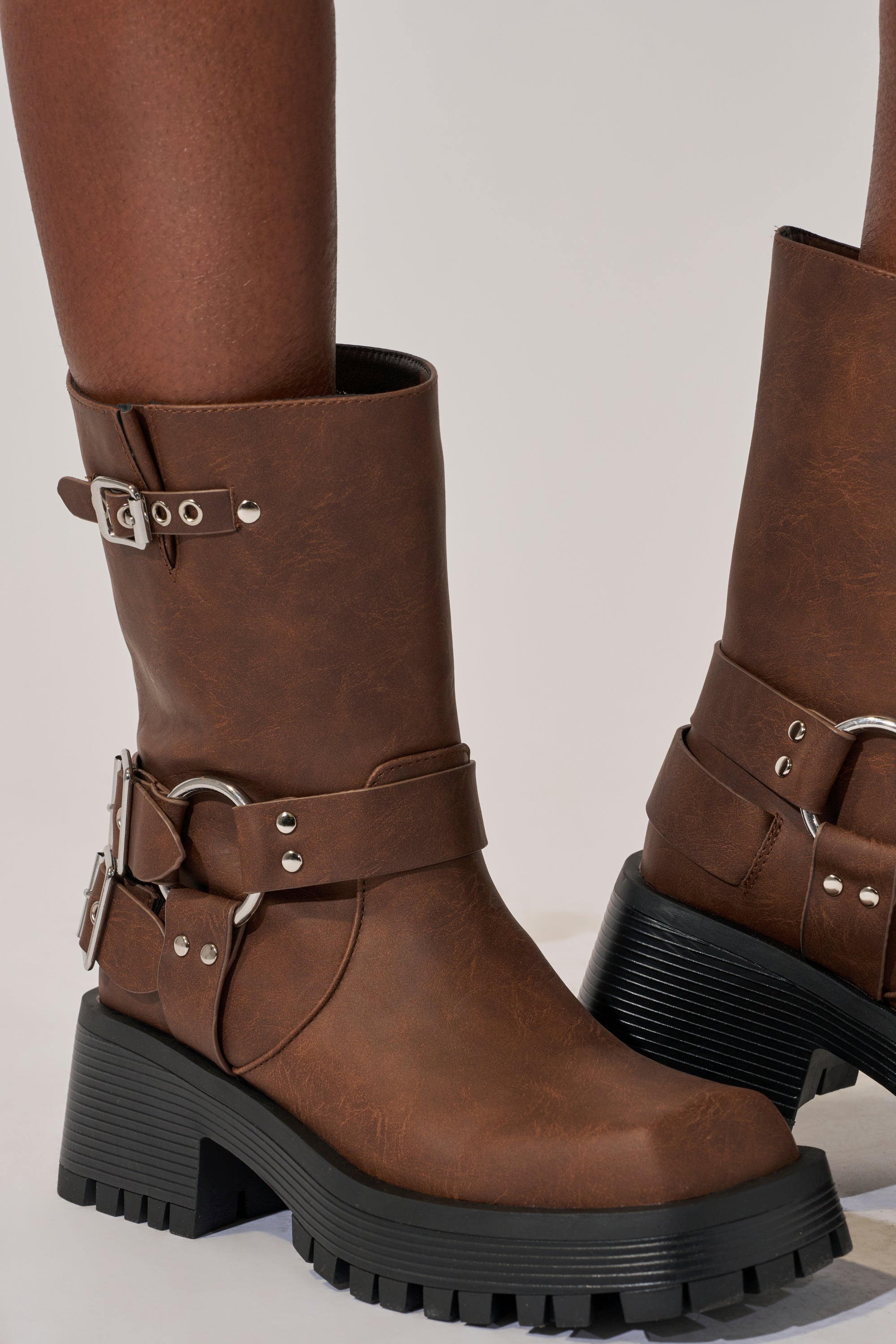 A close-up shows a person wearing AZALEA WANG BUCKED BROWN CHUNKY HEEL COMBAT BOOTIE, featuring mid-calf brown faux leather, silver buckles and straps, plus chunky black lug soles, set against a plain light background.