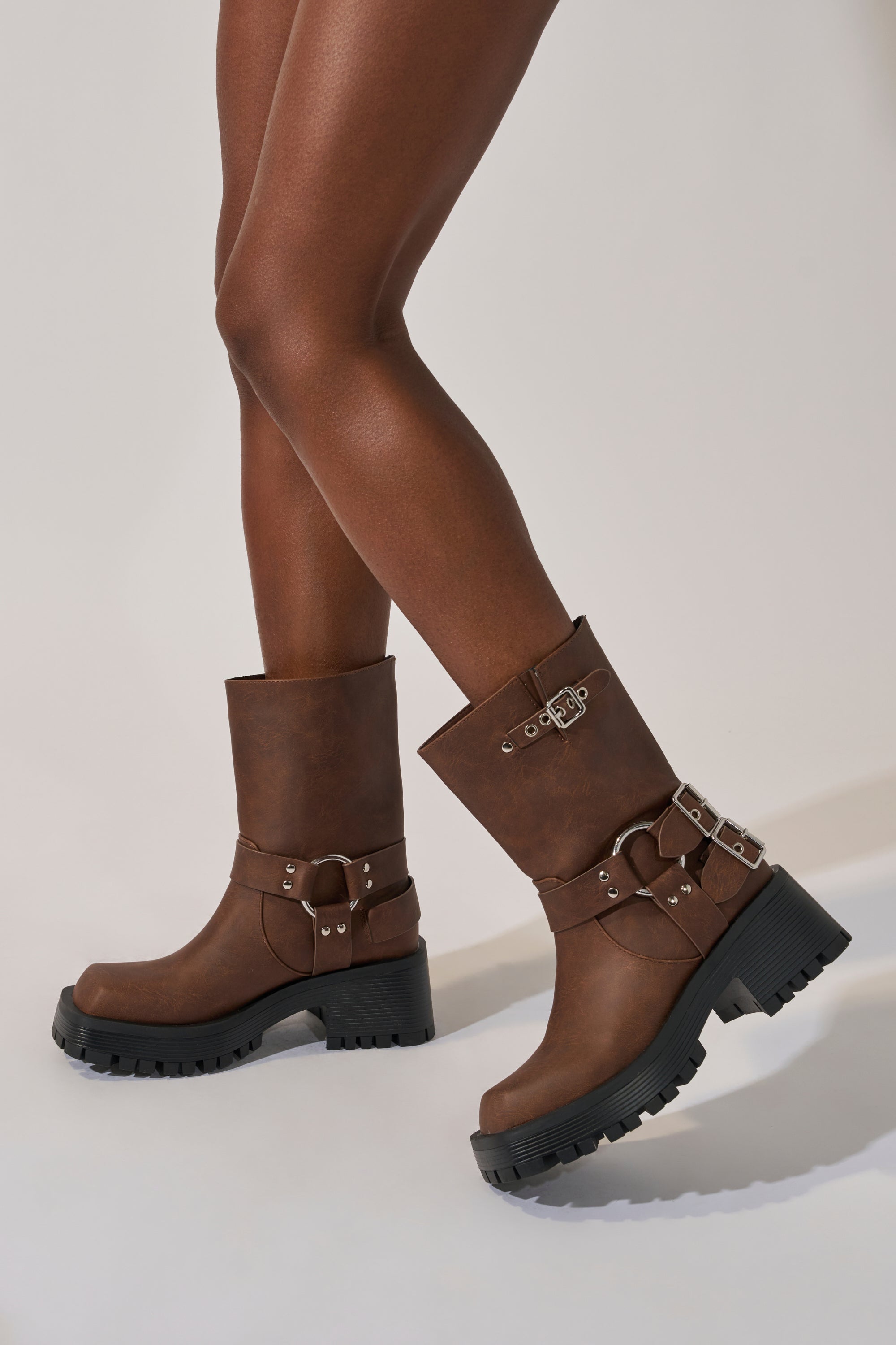 A person with medium-brown skin wears the AZALEA WANG BUCKED BROWN CHUNKY HEEL COMBAT BOOTIE, featuring brown faux leather, black chunky soles, and silver buckles, standing on a light background.