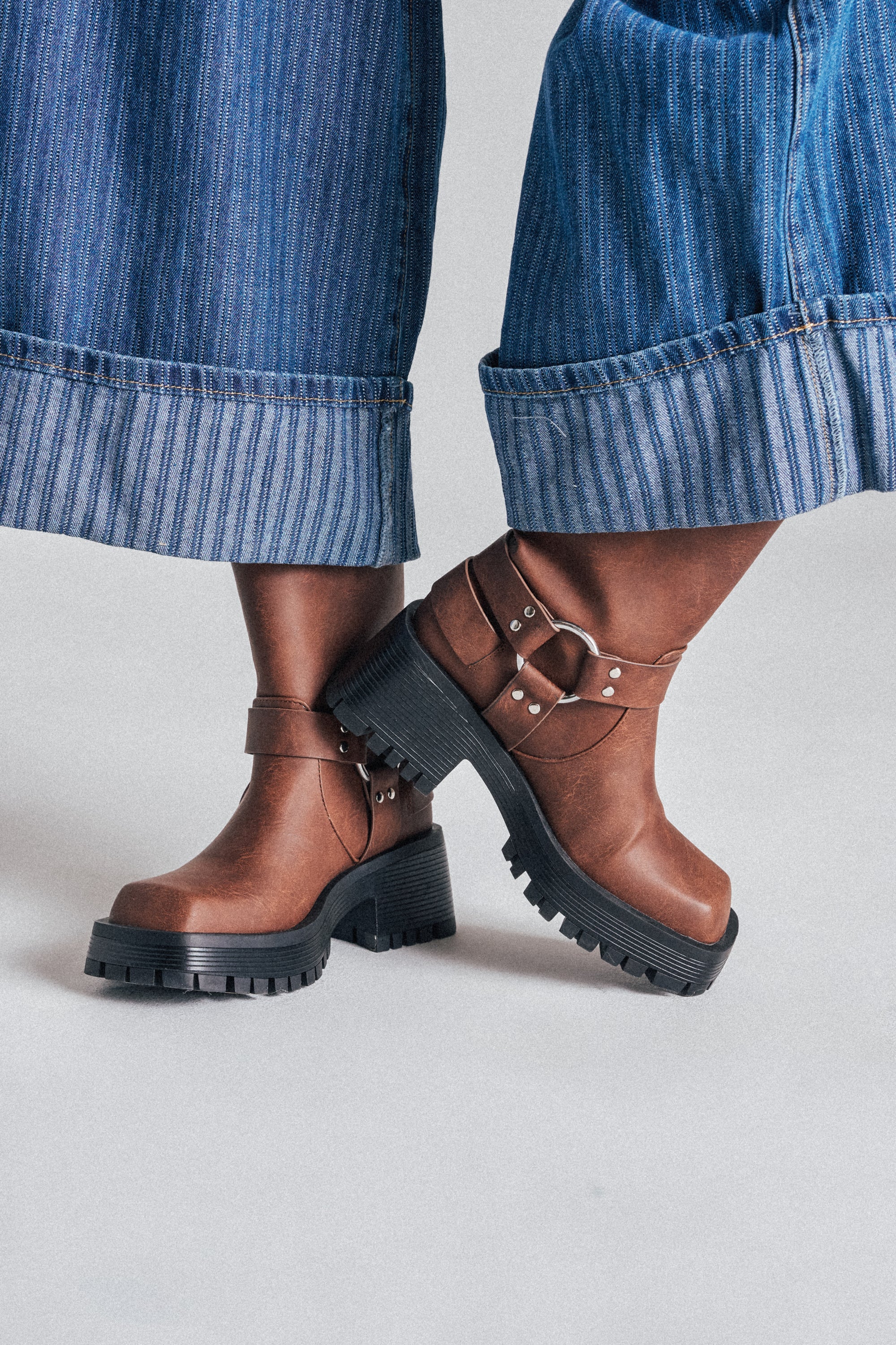 A person in wide-leg blue denim jeans wears AZALEA WANG BUCKED BROWN CHUNKY HEEL COMBAT BOOTIE, standing with one foot crossed in front of the other. 1