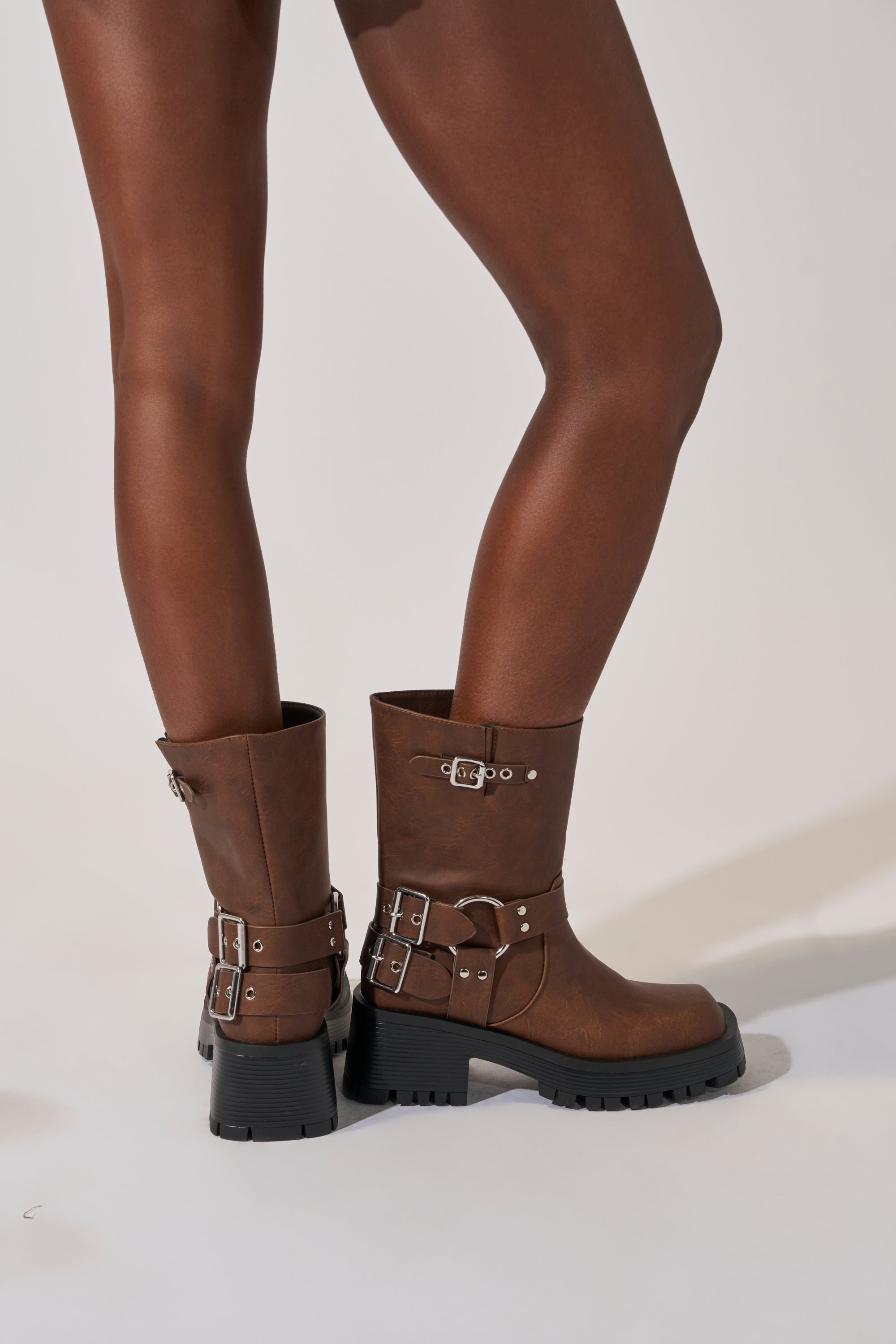 A person with dark skin models the AZALEA WANG BUCKED BROWN CHUNKY HEEL COMBAT BOOTIE, featuring brown mid-calf design, chunky black soles, and multiple silver buckles, set against a light background.