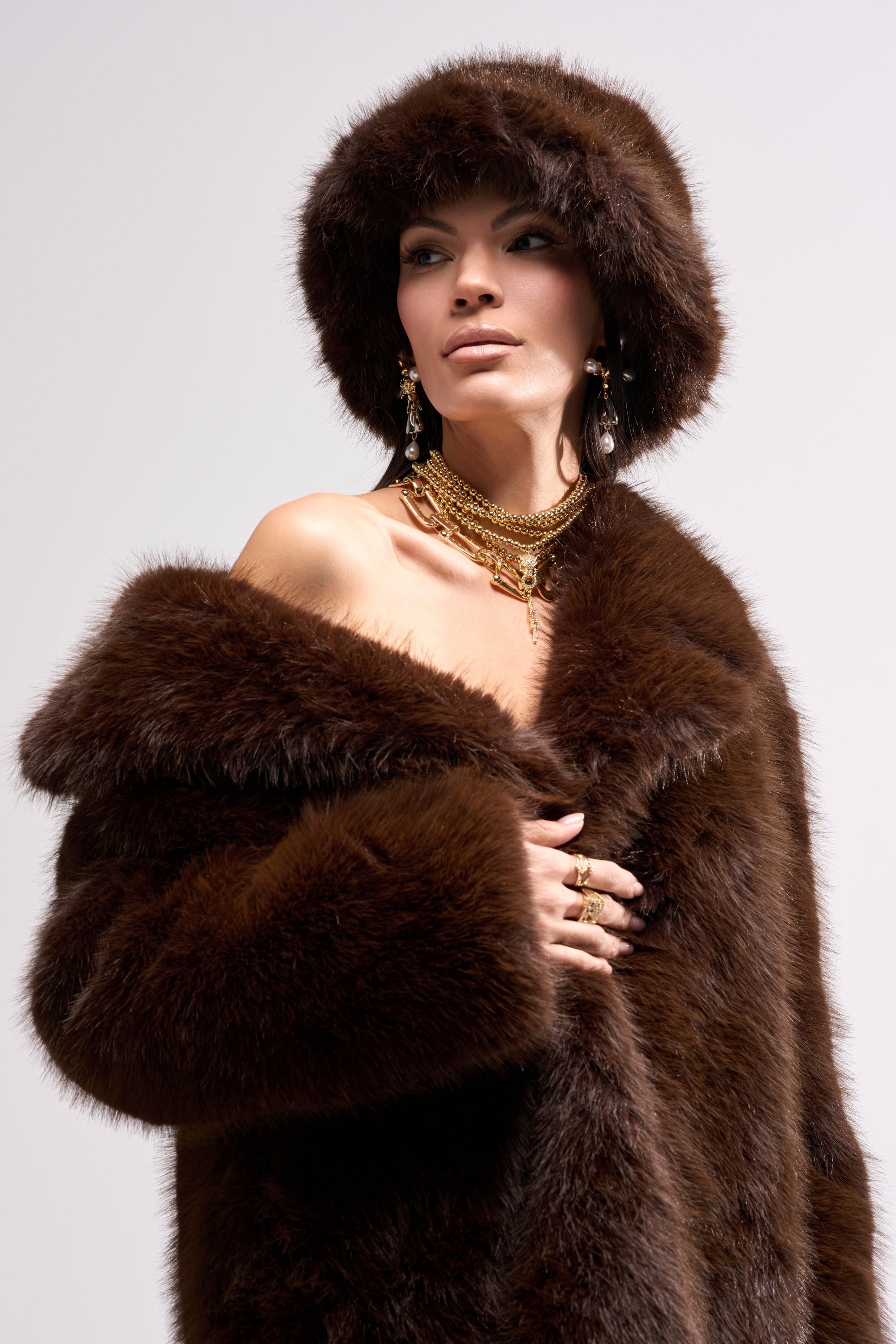 A woman confidently models the SO RICH FAUX FUR BUCKET HAT in brown, paired with a matching coat and gold jewelry against a light background—an effortlessly chic winter statement accessory.
