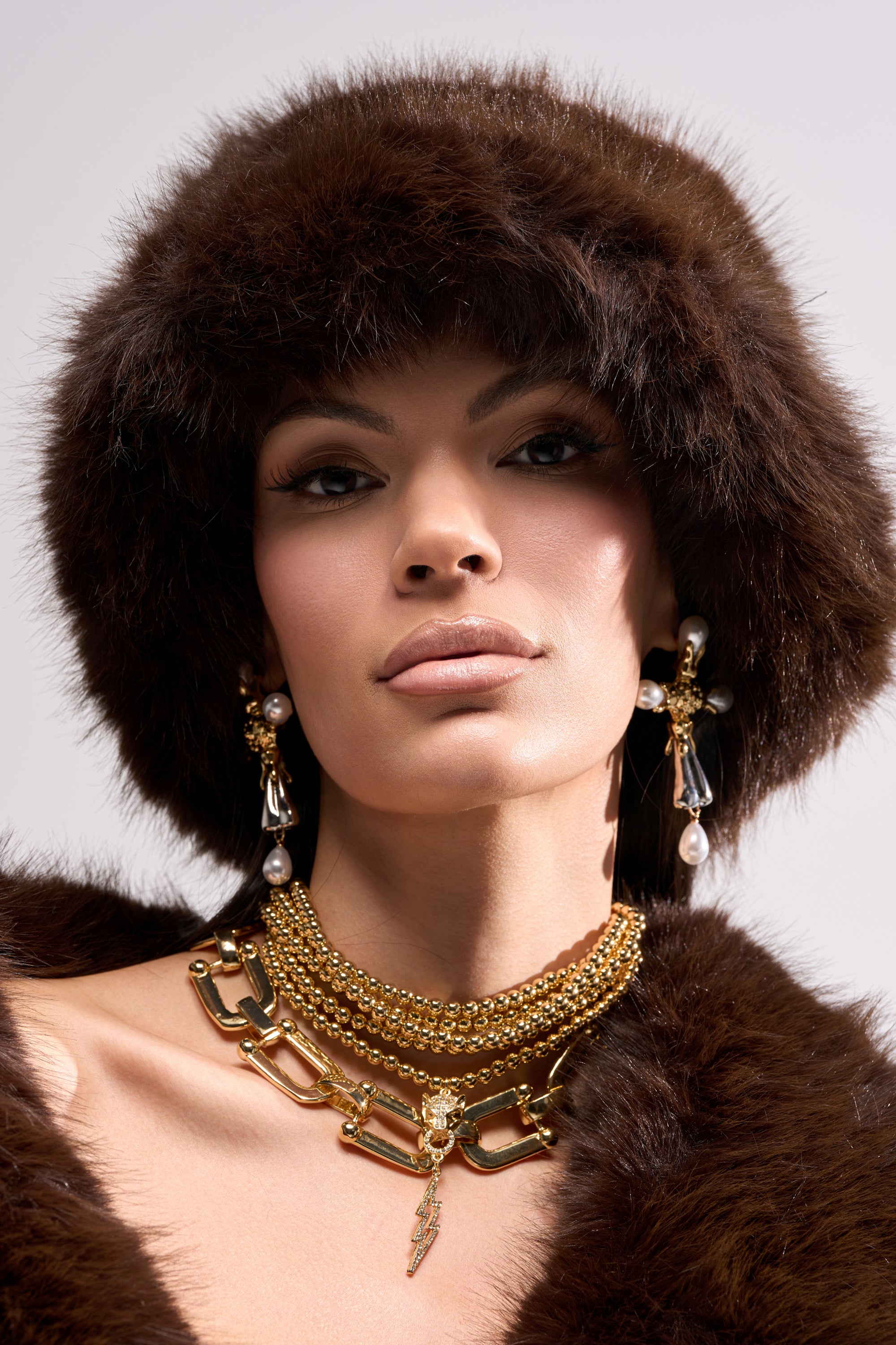A woman models the SO RICH FAUX FUR BUCKET HAT in dark brown with a matching coat, layered gold statement necklaces, and pearl and crystal drop earrings, all set against a simple light backdrop.