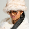 Wearing a stylish accessory ensemble, the person sports the TOASTIE FAUX FUR BUCKET HAT in beige alongside a cozy faux fur coat, paired perfectly with oversized round sunglasses and hoop earrings. Their long, dark hair falls gracefully against the simple background.