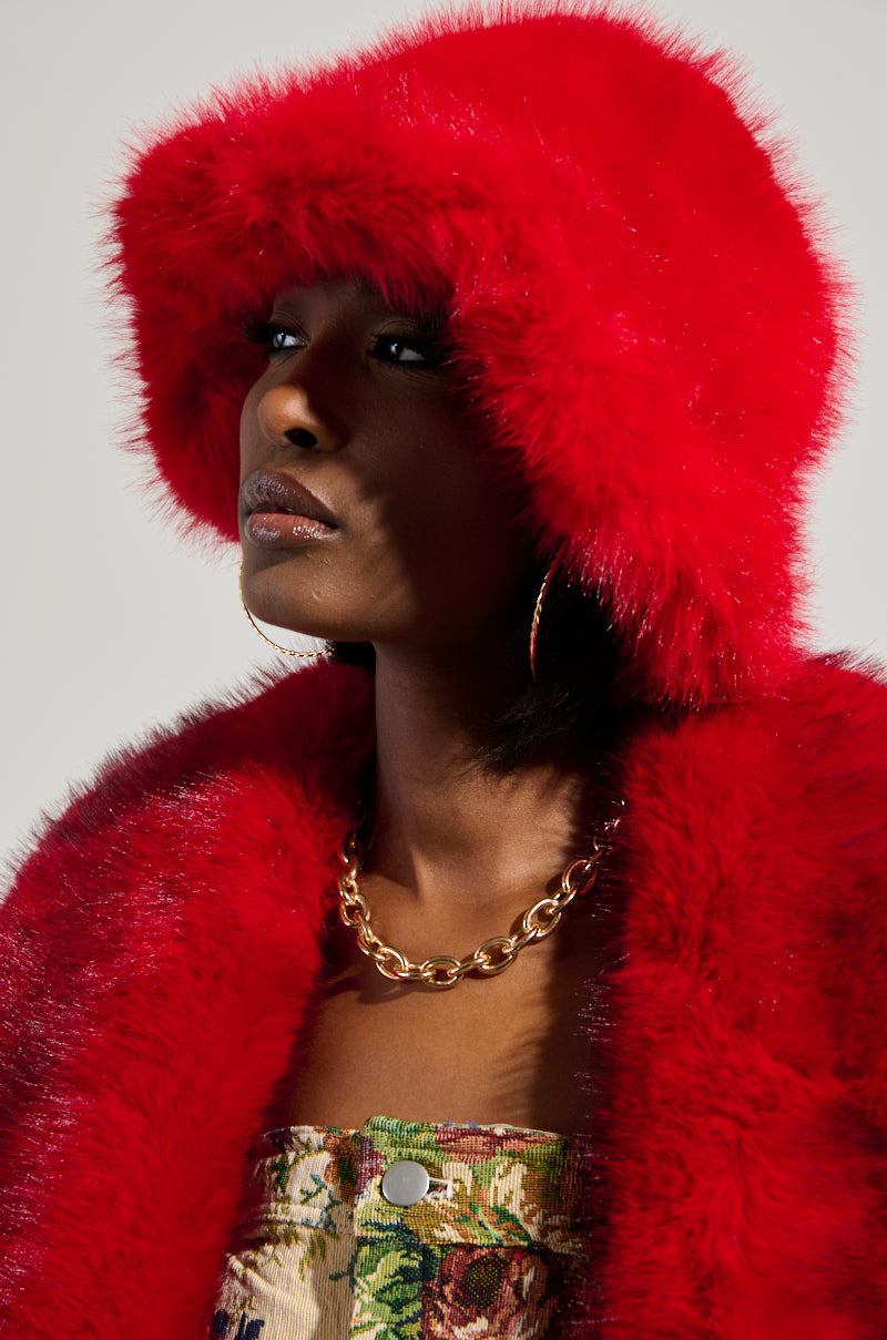 A person dressed in the LOOK AT ME NOW FAUX FUR HAT, boasting a vivid red hue perfect for cold weather and paired with a matching coat. They accessorize with large hoop earrings and a chunky gold chain necklace, while wearing a colorful patterned top against a plain background. 0