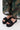 Close-up of a person wearing BUDZA BLACK FLATFORM SANDAL with an open almond toe and sheer, patterned green, gold, and black pants, standing on a light-colored floor.