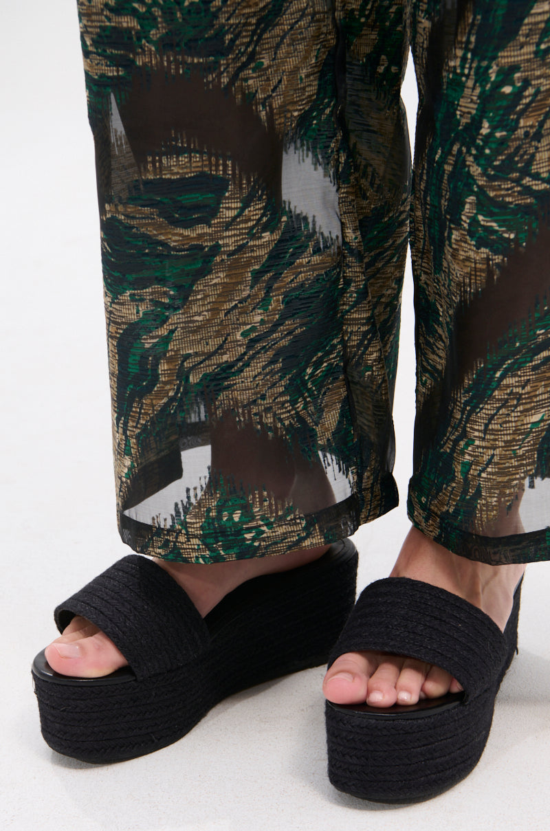 A close-up of a person wearing BUDZA BLACK FLATFORM SANDAL with crisscross straps and an open almond toe, styled with loose patterned green, brown, and beige pants against a plain light background.