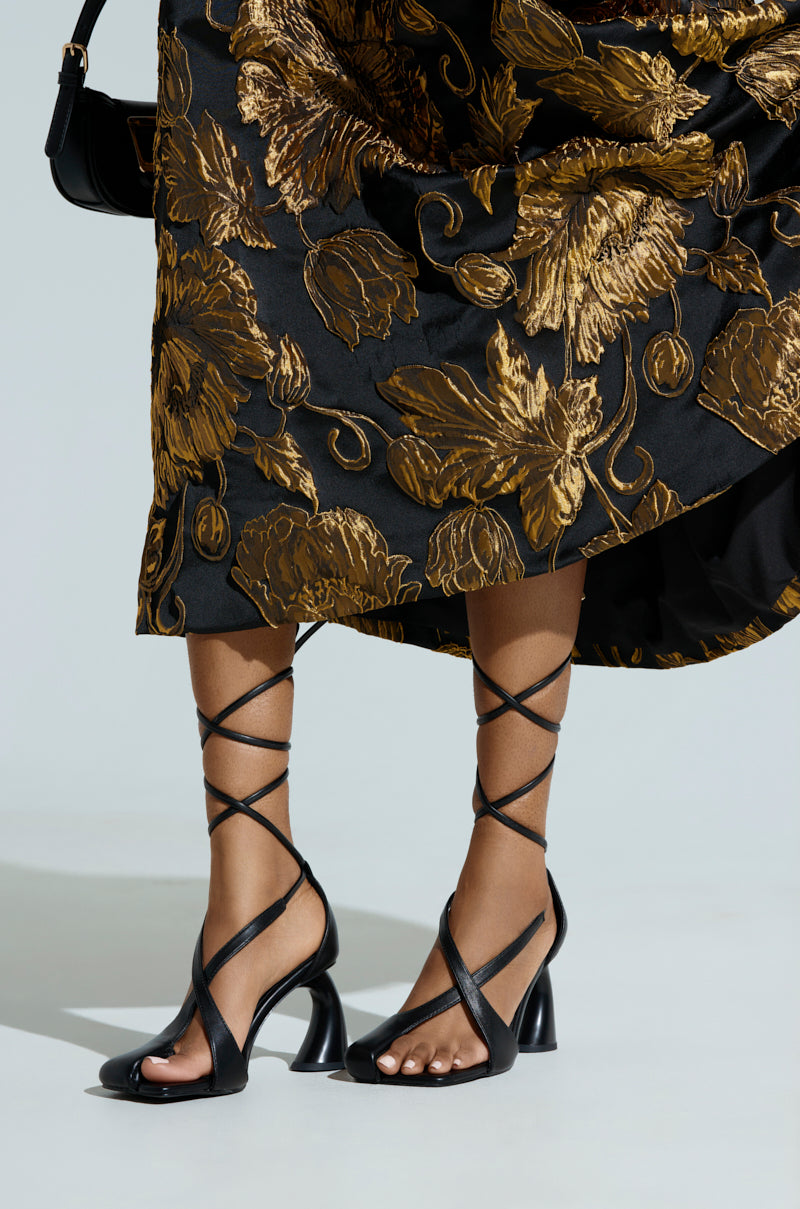 A close-up captures a person in AZALEA WANG BUIKY BLACK PUMP high-heeled sandals with sculpted heels and crisscross straps, paired with a golden floral patterned black skirt. They're holding a small black handbag against a plain light background.