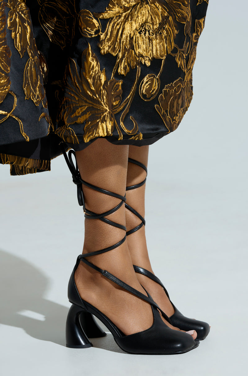 A close-up features someone wearing the AZALEA WANG BUIKY BLACK PUMP, showcasing its crisscross ankle straps and sculpted block heels. The faux leather complements a black skirt with large gold floral patterns, embracing the balletcore trend against a light gray background.