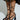 Close-up of a person wearing AZALEA WANG BUIKY BLACK PUMPs with strappy high-heel and intricate lace-up extending up the calves. Crafted in faux leather, they pair elegantly with a black garment adorned in gold floral patterns against a light, neutral background.