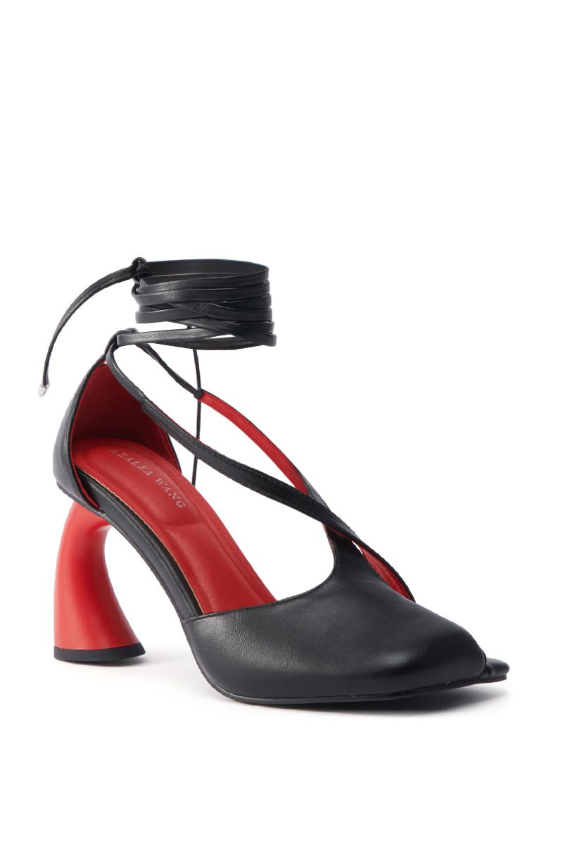 The AZALEA WANG BUIKY BLACK AND RED PUMP features a chunky sculptural heel, faux leather upper, red insole, cross-over straps, and an ankle wrap tie, all set against a white background.