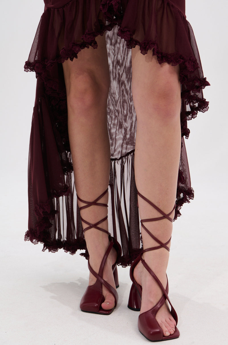 Someone stands on a light background wearing a burgundy ruffled high-low skirt and AZALEA WANG BUIKY BURGUNDY OPEN TOE PUMPS featuring crisscross straps that wrap around their calves.