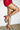 A person stands against a white wall with one leg bent, wearing a short red skirt and the AZALEA WANG BUIKY SNAKE CONTRAST CURVED HEEL OPEN TOE PUMP, featuring lace-up straps and red soles.
