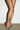 Someone with crossed legs stands on a concrete floor against a white brick wall, wearing the AZALEA WANG BUIKY SNAKE CONTRAST CURVED HEEL OPEN TOE PUMP featuring a snake print and red accents.