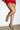 A woman in a short red skirt stands against a white wall, wearing the AZALEA WANG BUIKY SNAKE CONTRAST CURVED HEEL OPEN TOE PUMP. Only her legs and part of the red skirt are visible.